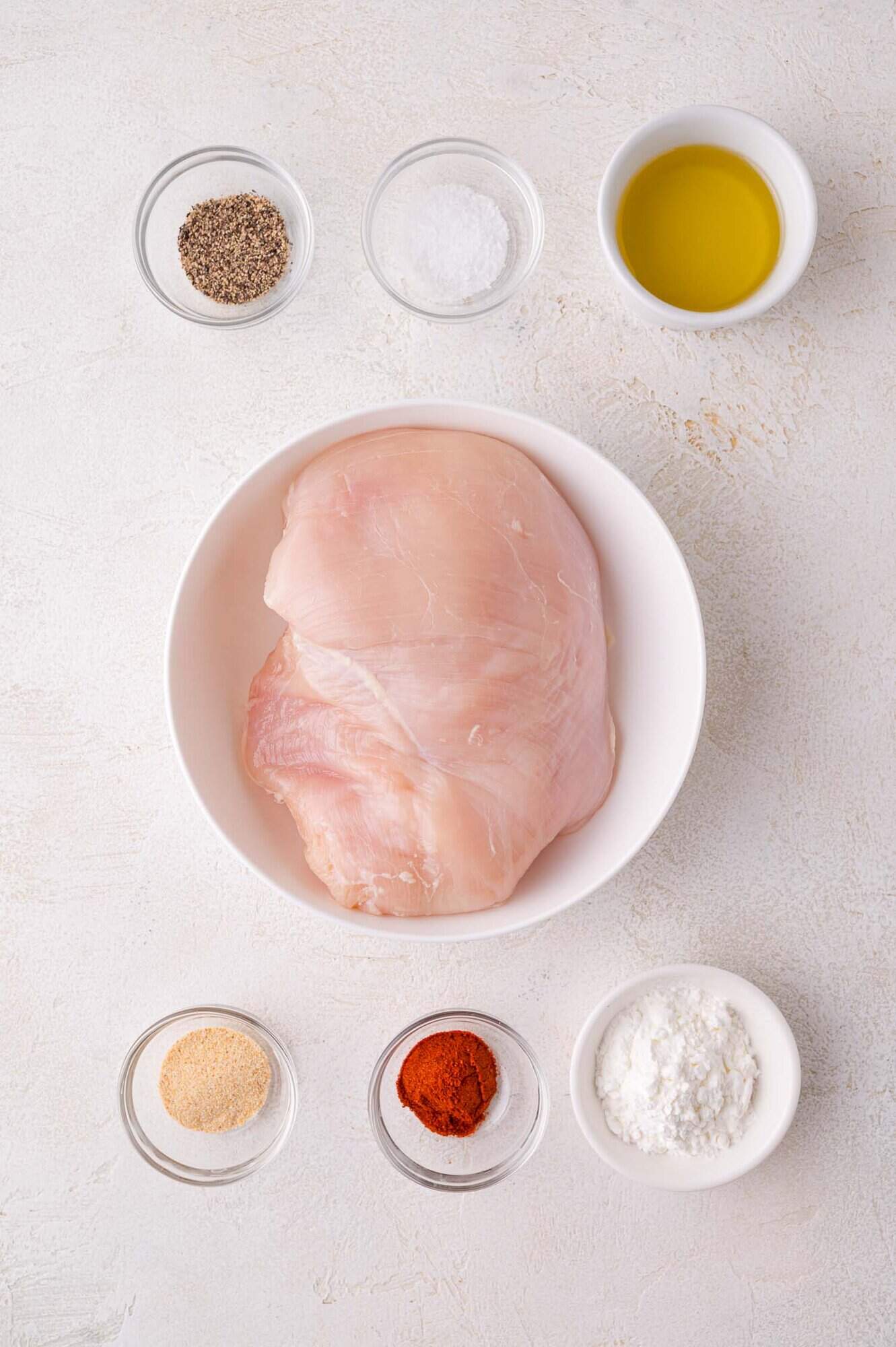 Raw chicken breast in a white bowl surrounded by small bowls containing black pepper, salt, olive oil, garlic powder, paprika, and cornstarch on a light surface.