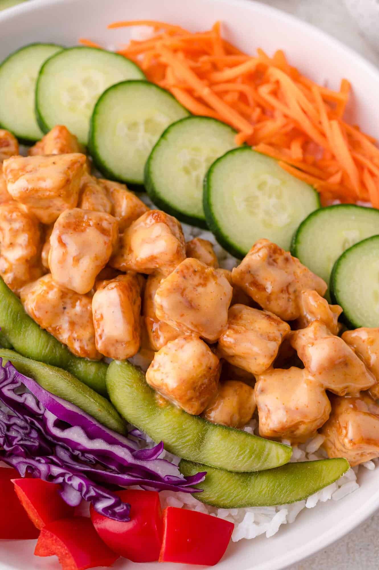 A close-up of a bowl with white rice, cooked chicken pieces in bang bang sauce, sliced cucumbers, shredded carrots, snap peas, shredded purple cabbage, and diced red bell pepper.