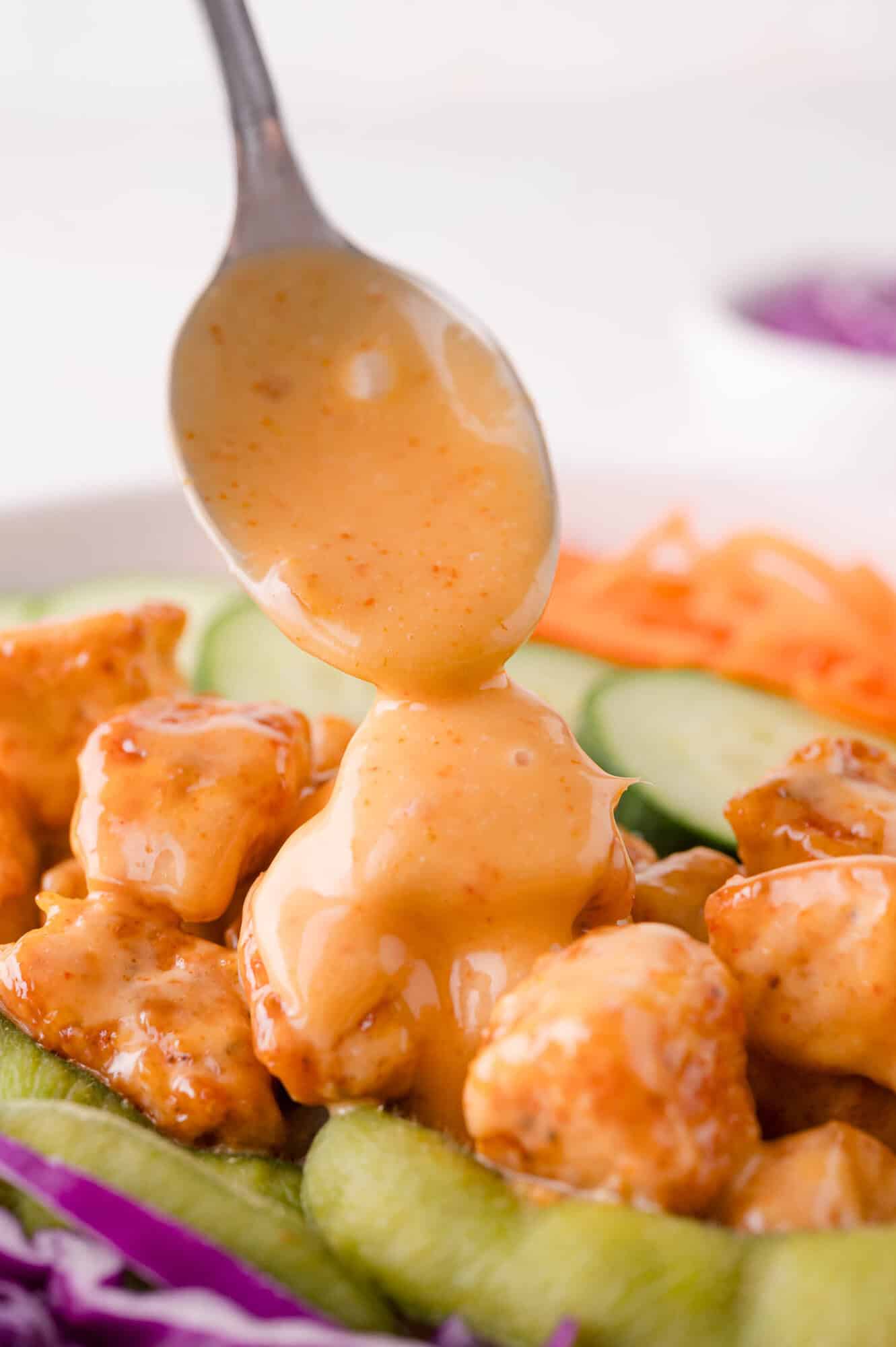 A spoon drizzling creamy bang bang sauce over pieces of cooked chicken, with sliced cucumber, shredded carrots, and cabbage in the background.