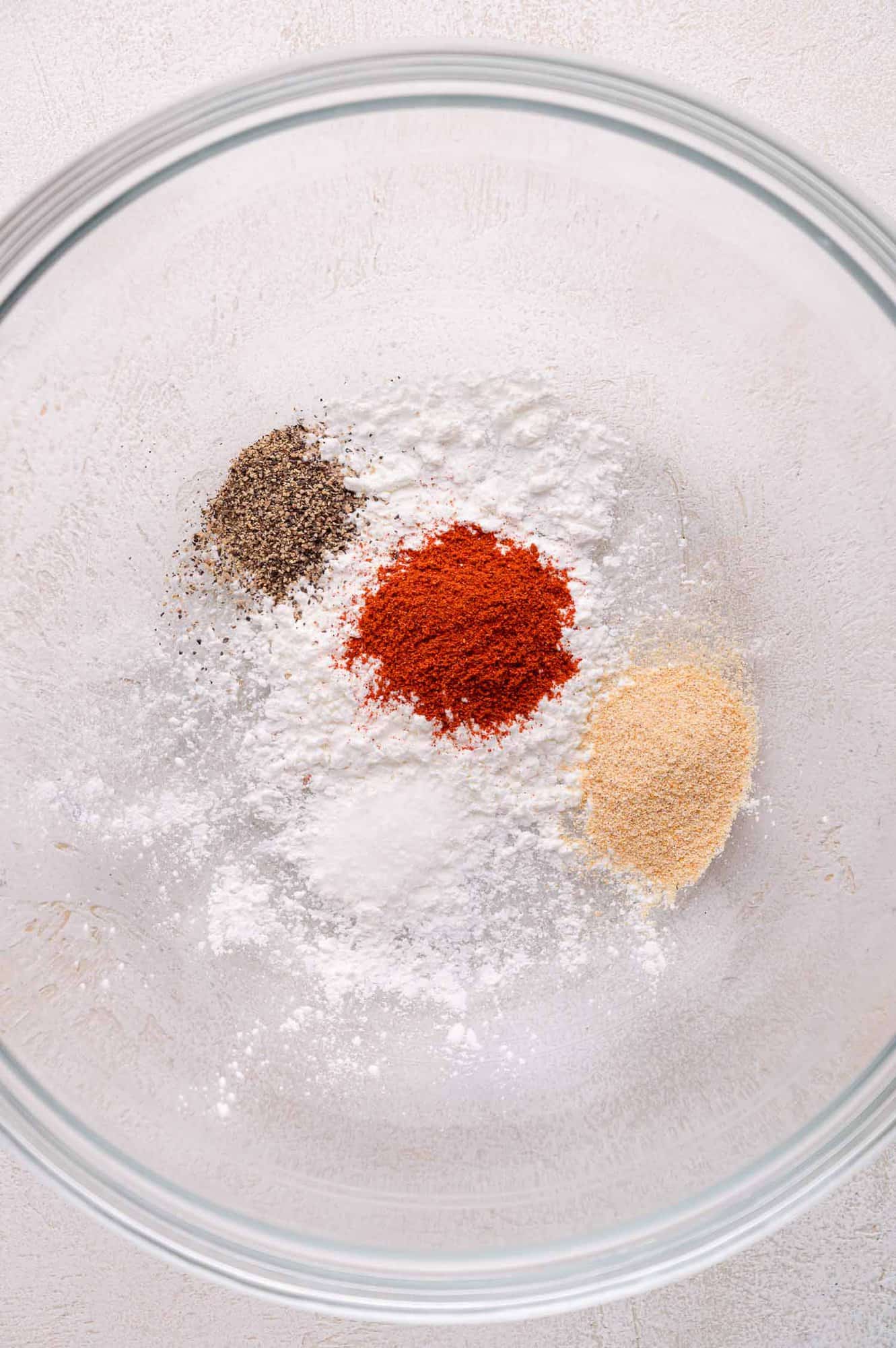 A glass bowl containing separate piles of ground black pepper, paprika, garlic powder, and cornstarch on a light surface.