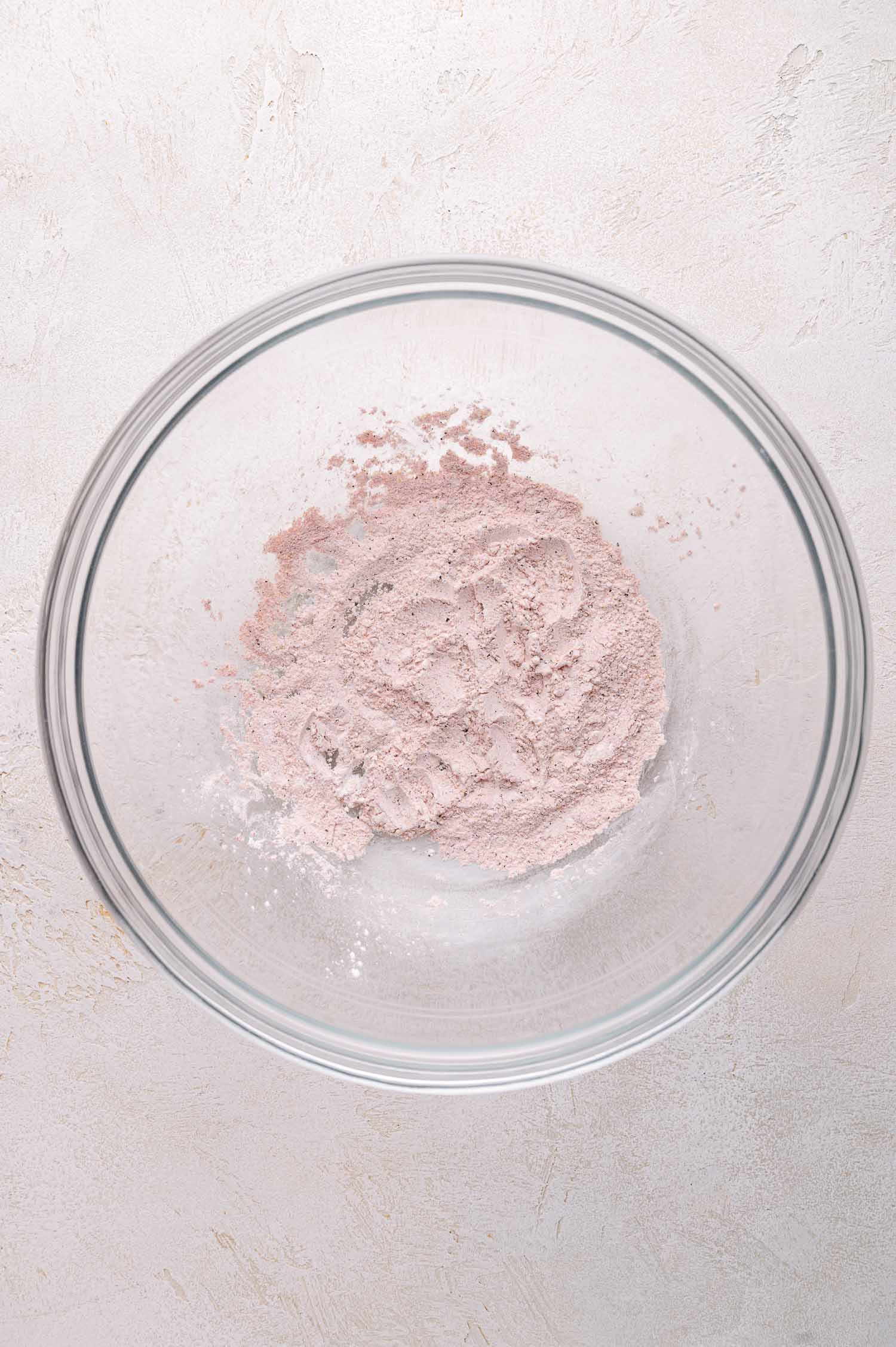 A glass mixing bowl containing a small amount of light pink powder sits on a textured, light-colored surface.