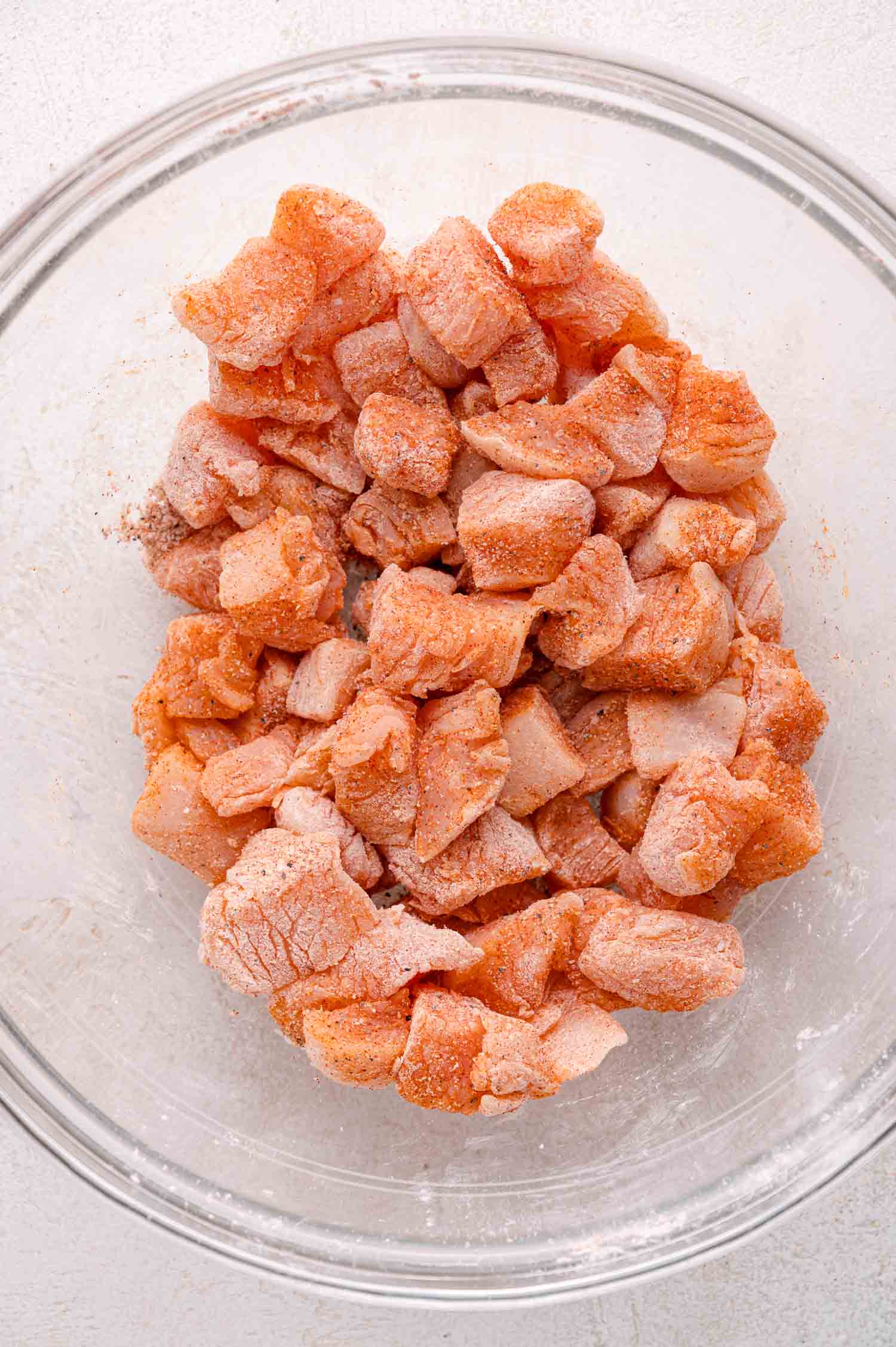 Raw, cubed chicken pieces coated with spices in a clear glass mixing bowl on a light-colored surface.