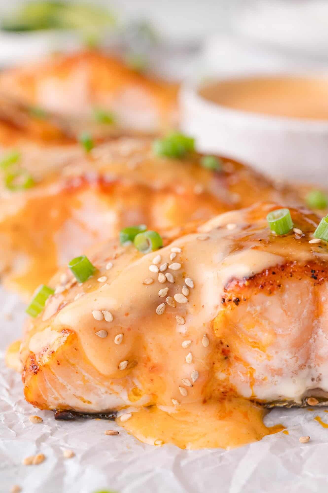 Close-up of cooked salmon fillets topped with creamy sauce, sesame seeds, and chopped green onions, served on parchment paper.