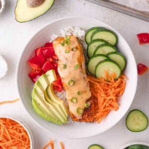 A bowl of white rice topped with bang bang salmon, sliced cucumber, avocado, diced tomatoes, shredded carrots, and a creamy sauce, garnished with green onions and sesame seeds.