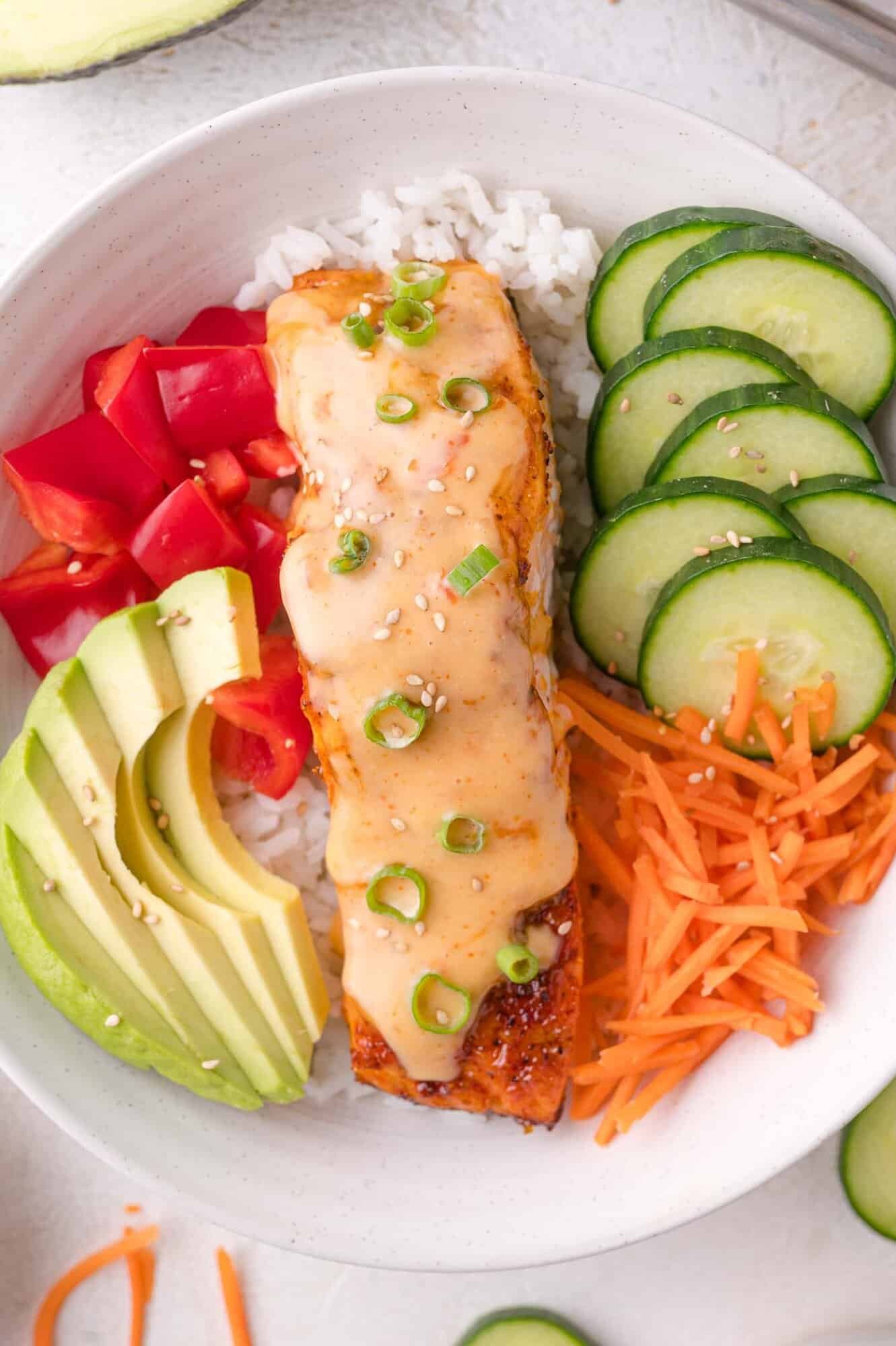 A bowl of white rice topped with a glazed salmon fillet, sliced cucumber, shredded carrots, avocado, chopped red bell pepper, and garnished with sesame seeds and green onion.