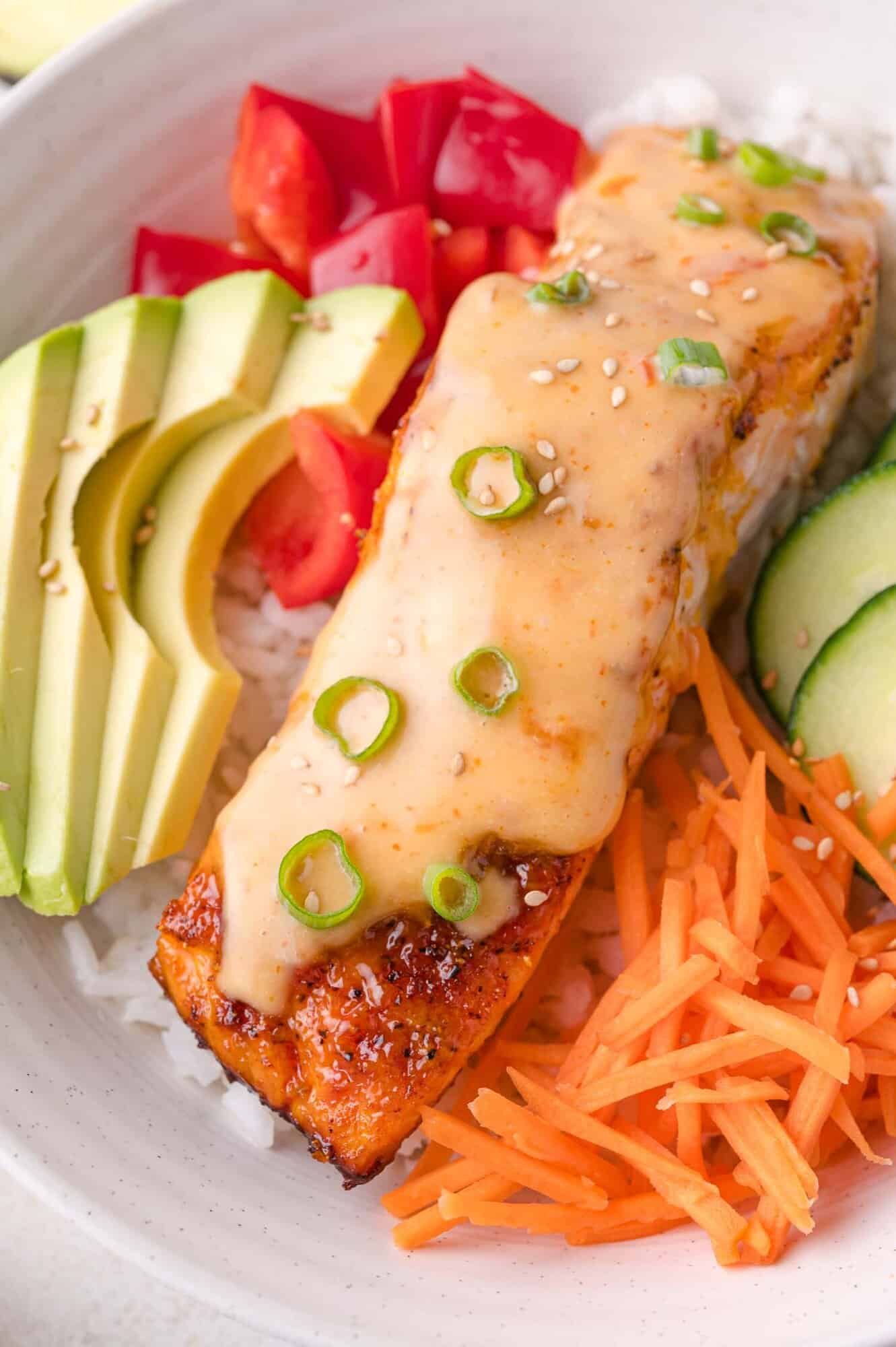 Bowl with rice, sliced avocado, tomatoes, cucumbers, shredded carrots, and a piece of bang bang salmon topped with sesame seeds and sliced green onions.