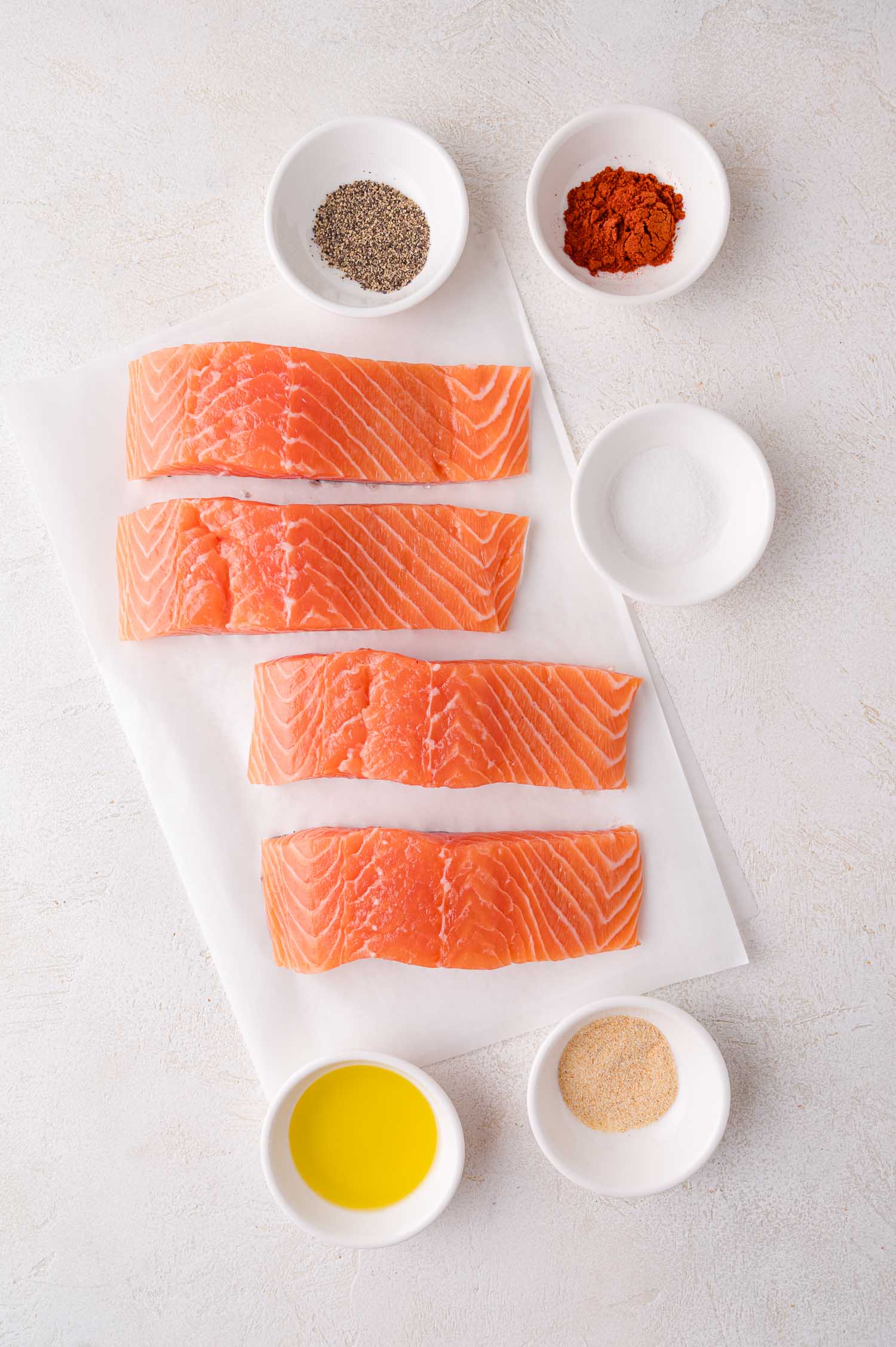 Four raw salmon fillets on parchment paper, surrounded by small white bowls containing pepper, paprika, salt, olive oil, and garlic powder.