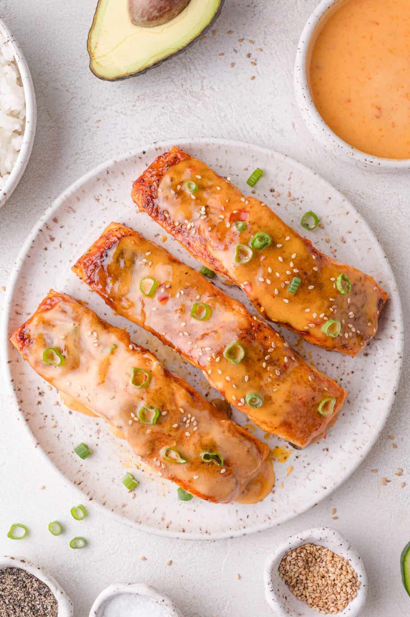 Three pieces of bang bang sauce glazed salmon topped with sliced green onions and sesame seeds are arranged on a speckled plate, with sauce, rice, seasonings, and half an avocado nearby.