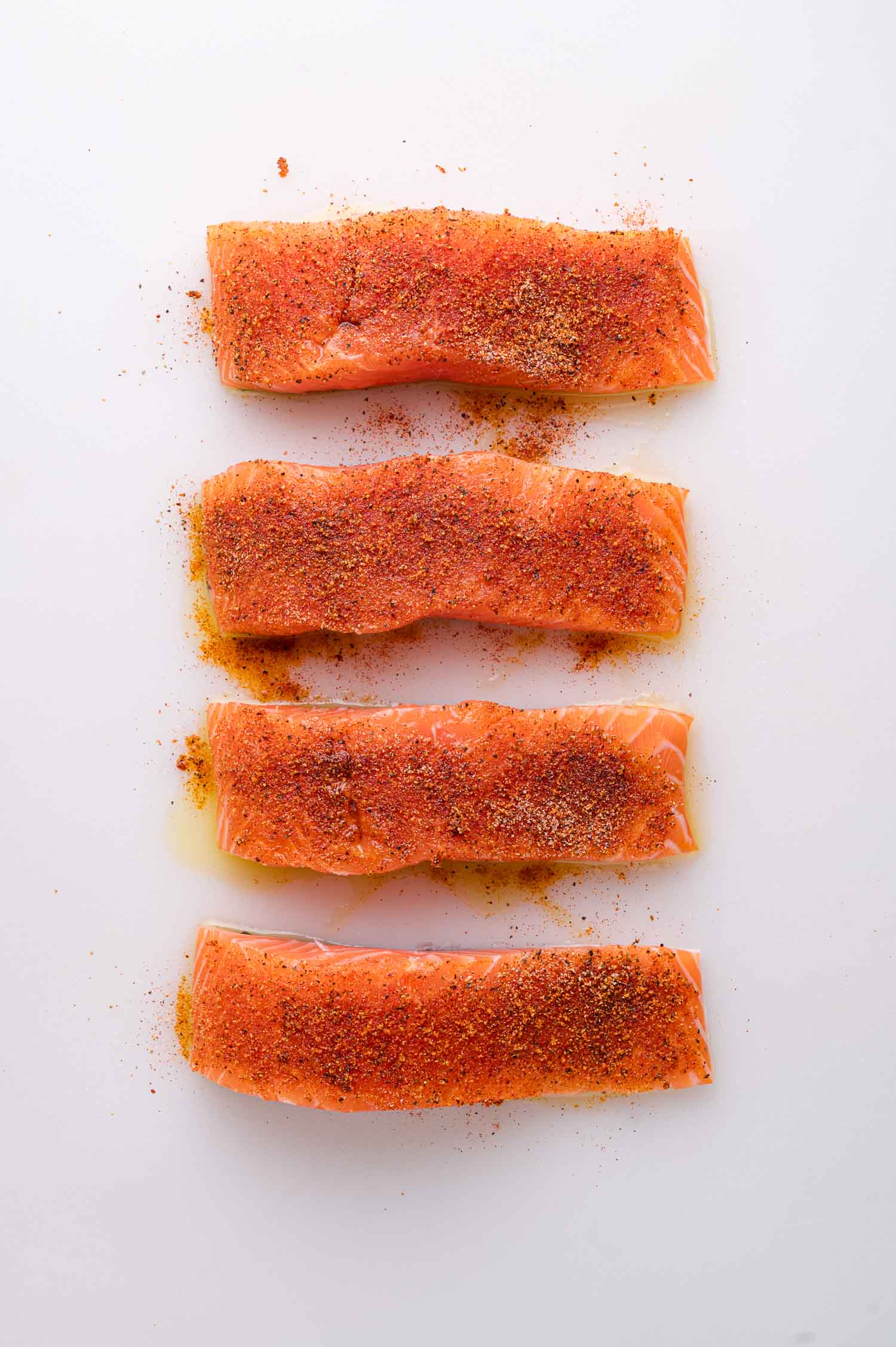 Four raw salmon fillets arranged vertically on a white surface, each topped with a mix of red and brown spices.