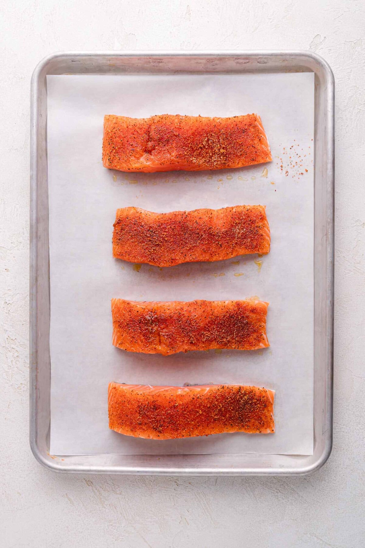 Four seasoned salmon fillets are arranged in a row on a parchment-lined baking sheet.