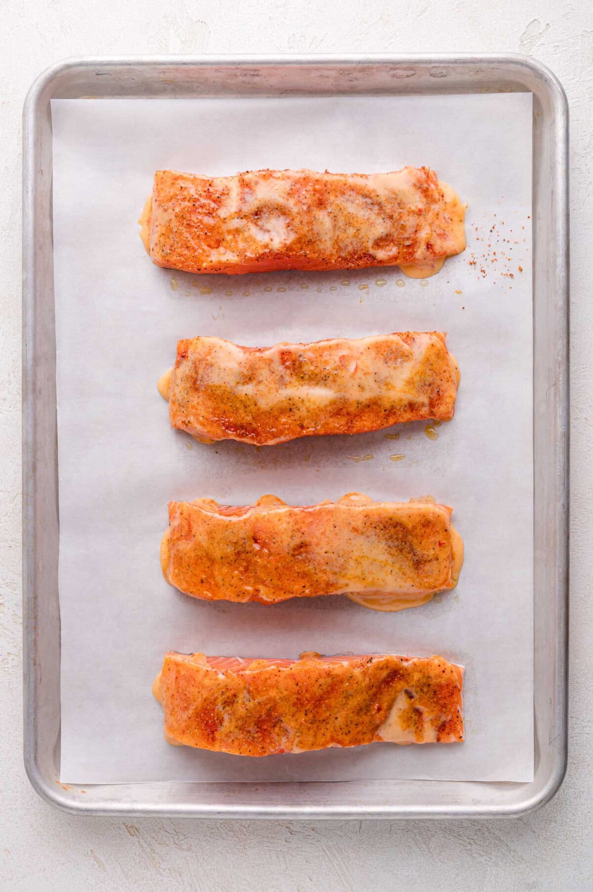 Four seasoned salmon fillets are arranged in a single row on a parchment-lined baking sheet, ready for cooking.