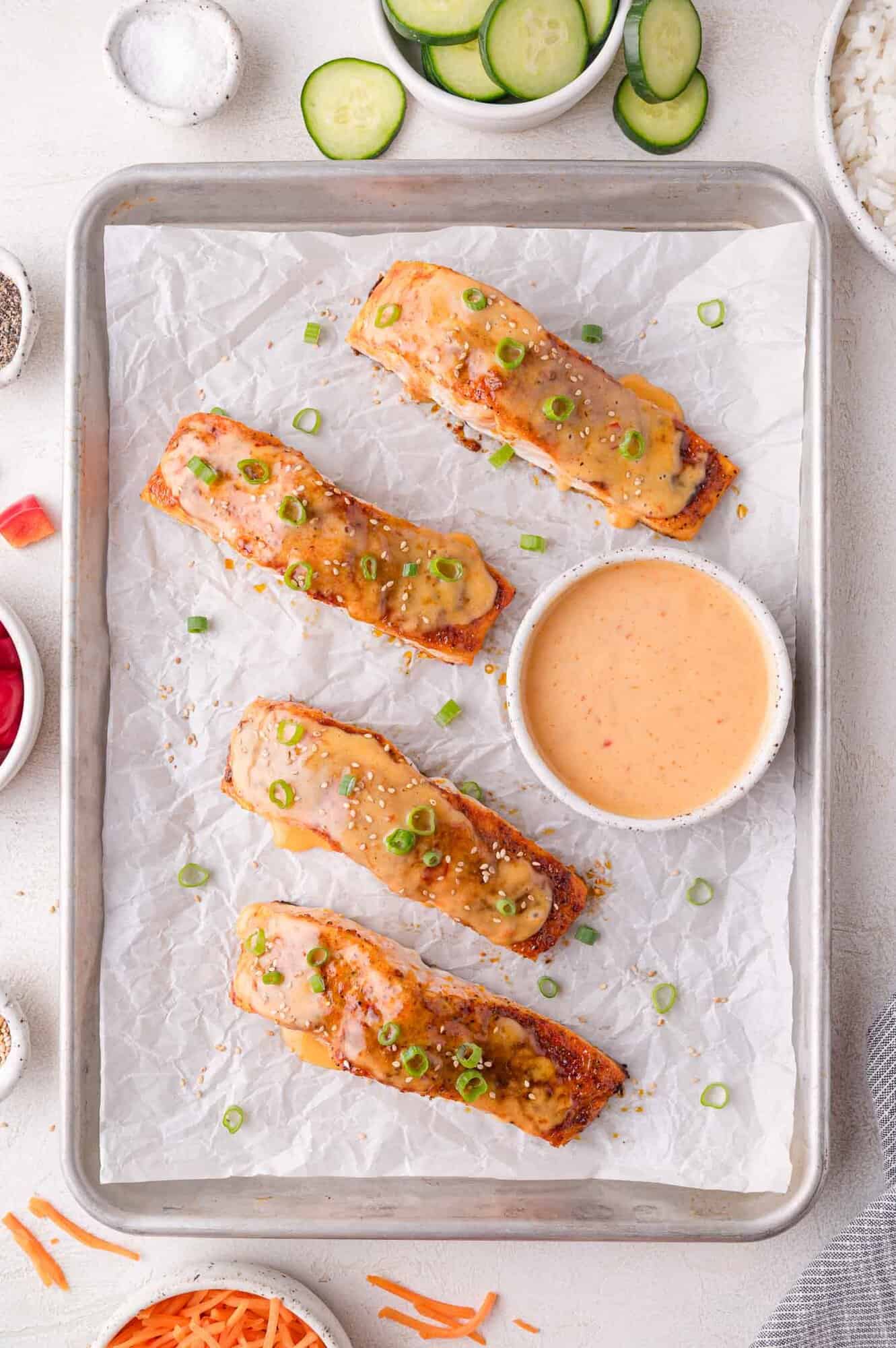 Four baked salmon fillets topped with sauce and chopped green onions on a parchment-lined baking sheet, with a bowl of dipping sauce in the center. Surrounding bowls contain sliced cucumbers and rice.
