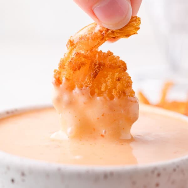 A hand dips a breaded, fried shrimp into a creamy orange bang bang sauce in a white bowl.