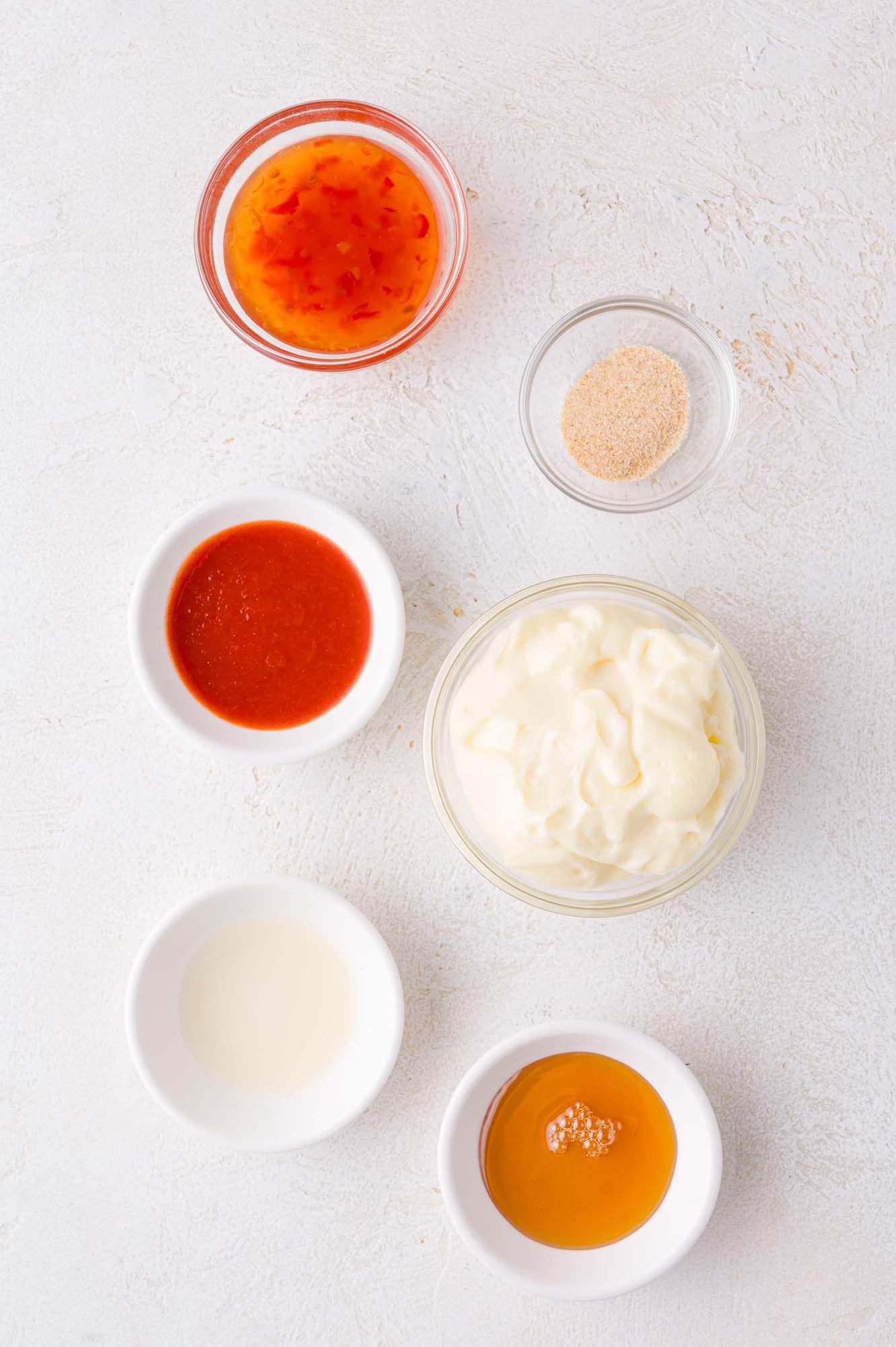 Six small bowls containing mayonnaise, sweet chili sauce, sriracha, garlic powder, vinegar, and honey are arranged on a white surface.