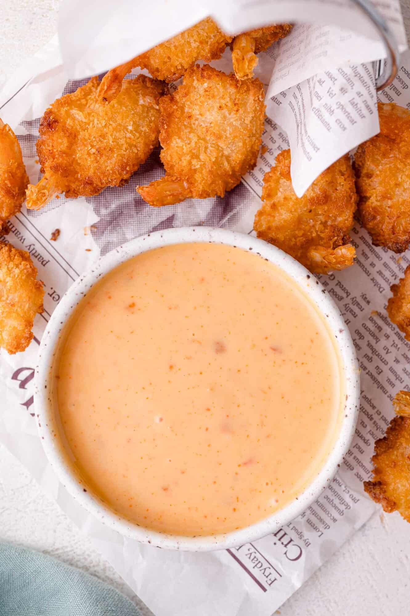 A bowl of creamy bang bang dipping sauce sits on newspaper next to several pieces of fried breaded shrimp.