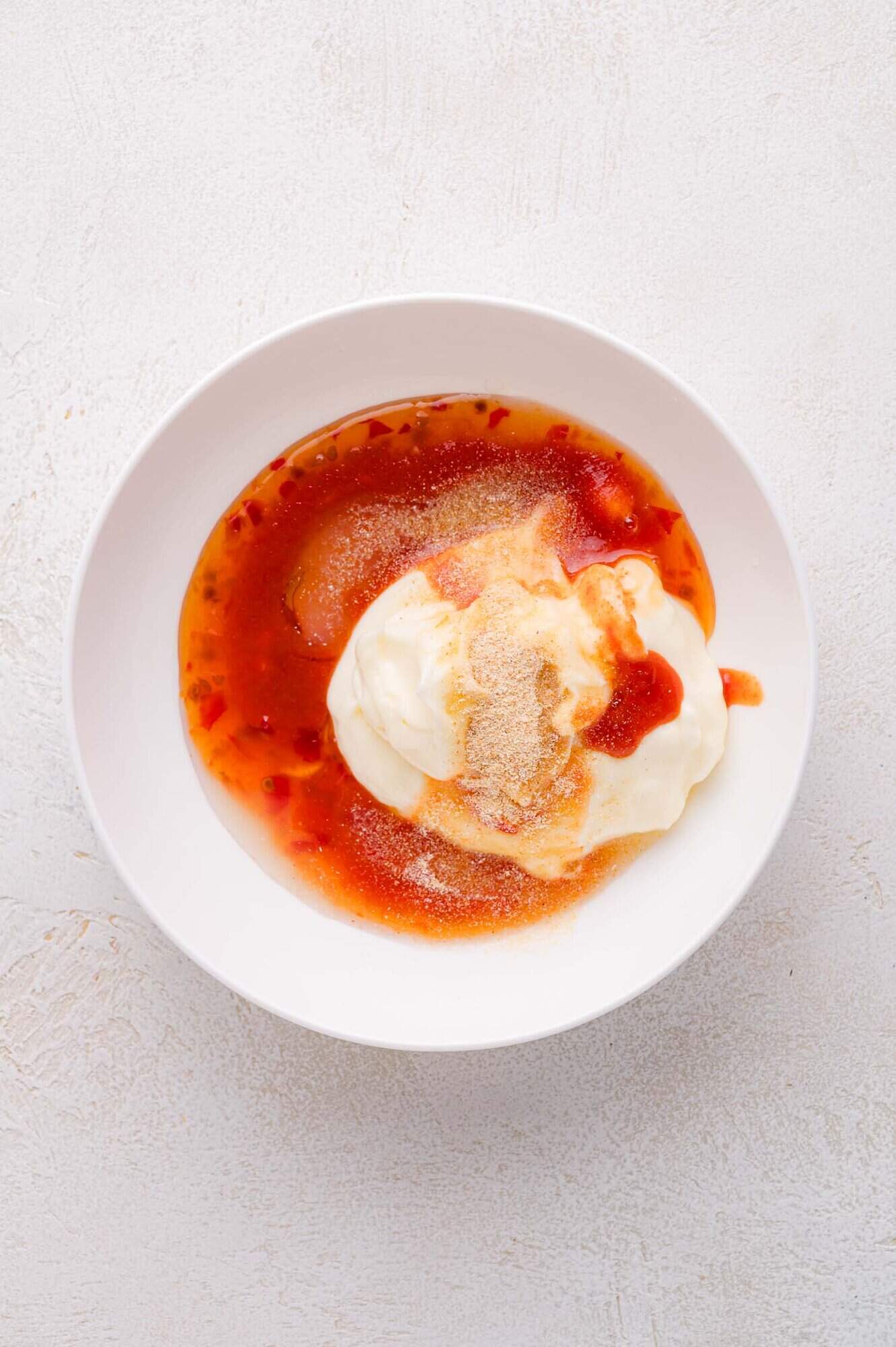 A white bowl contains mayonnaise, sweet chili sauce, and seasonings on a light surface.