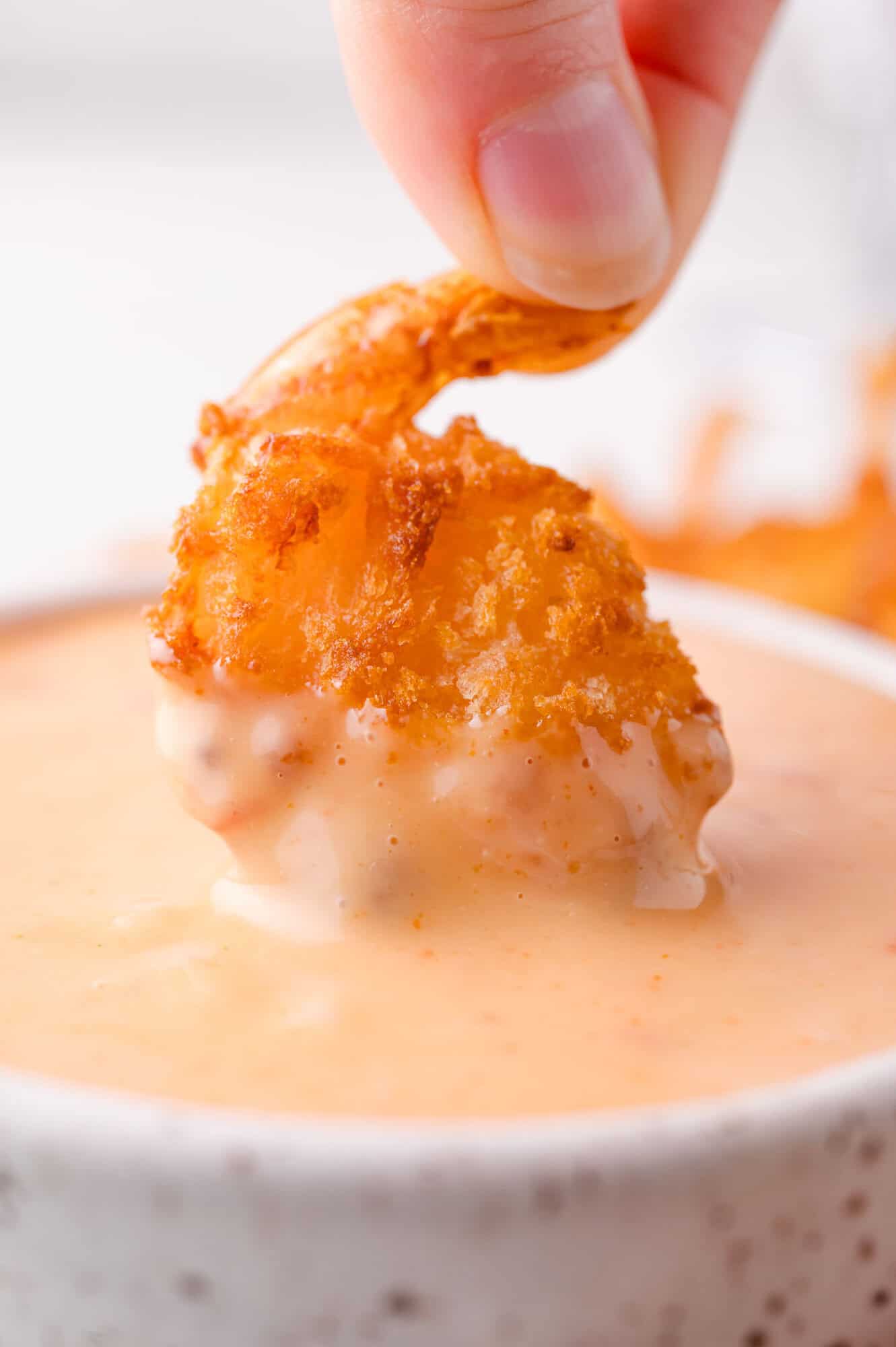 A hand dips a piece of crispy fried shrimp into a creamy, orange-colored sauce in a white bowl.