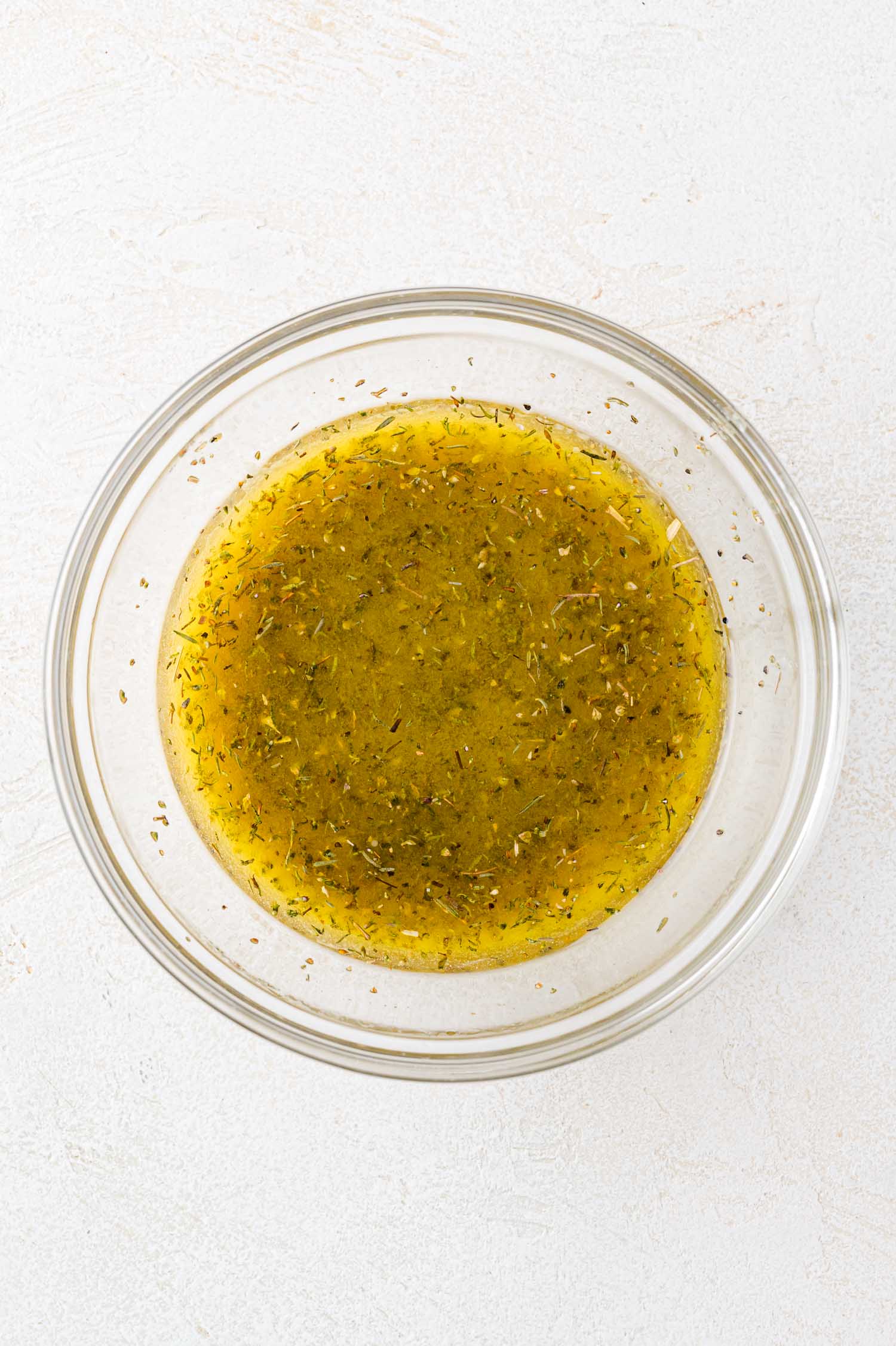 A clear glass bowl filled with yellowish vinaigrette dressing containing visible herbs, placed on a light-colored surface.