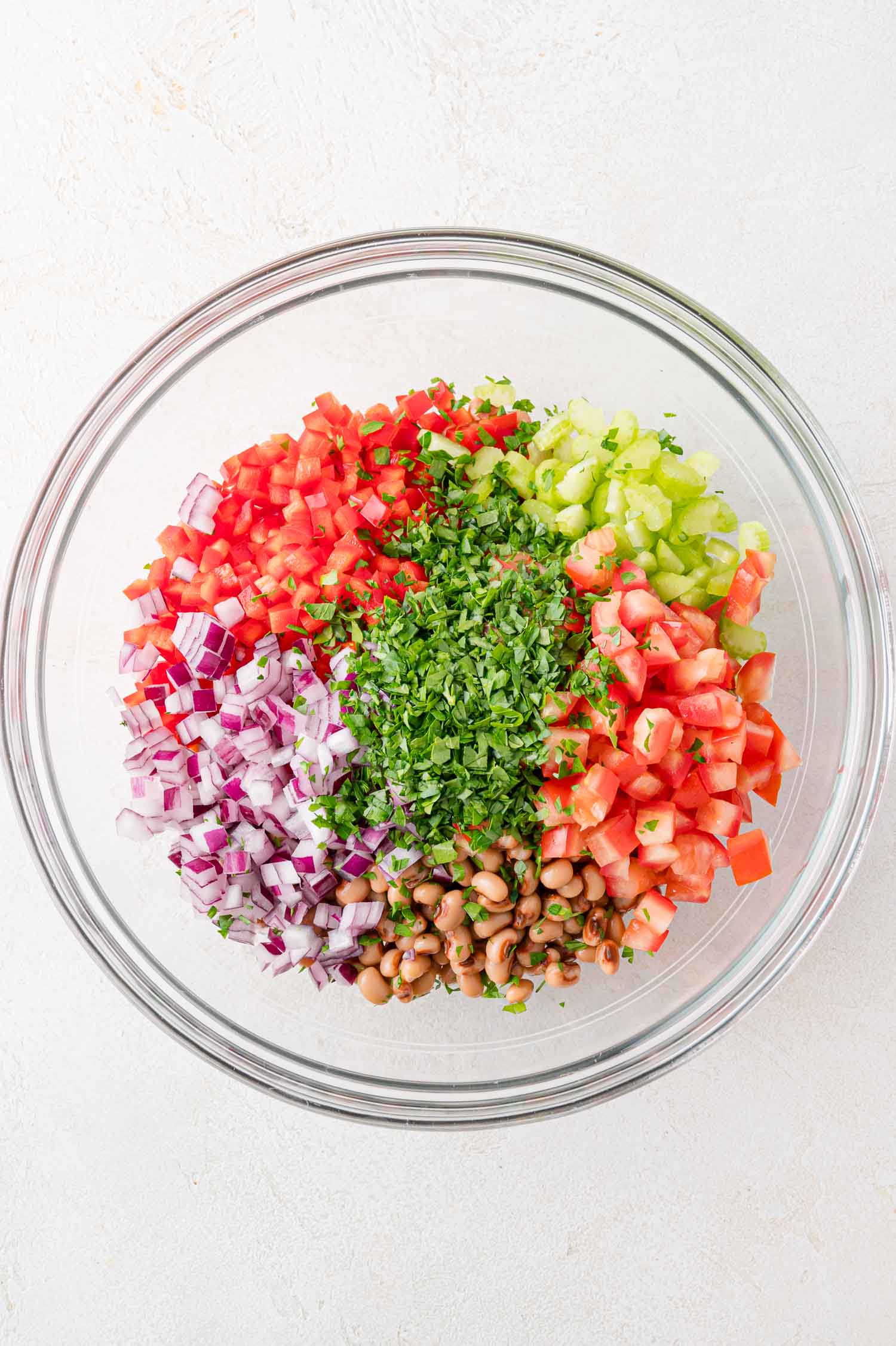 A glass bowl containing diced red onions, tomatoes, celery, red bell pepper, chopped parsley, and black-eyed peas, arranged in sections on a white surface.