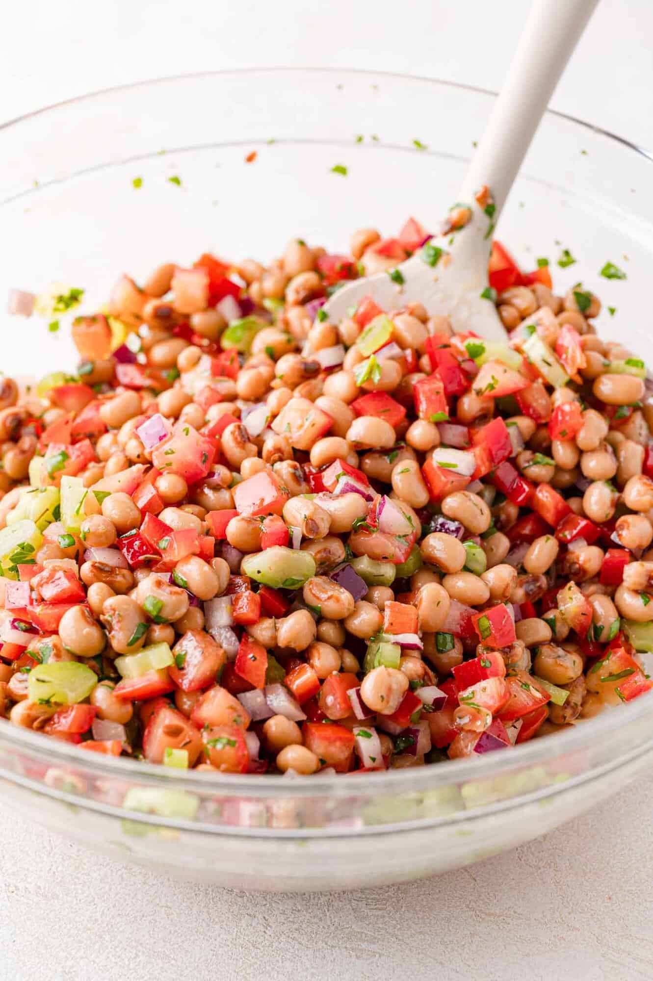 A glass bowl filled with a bean salad containing black-eyed peas, diced tomatoes, red onion, celery, and herbs, with a white spoon for mixing.
