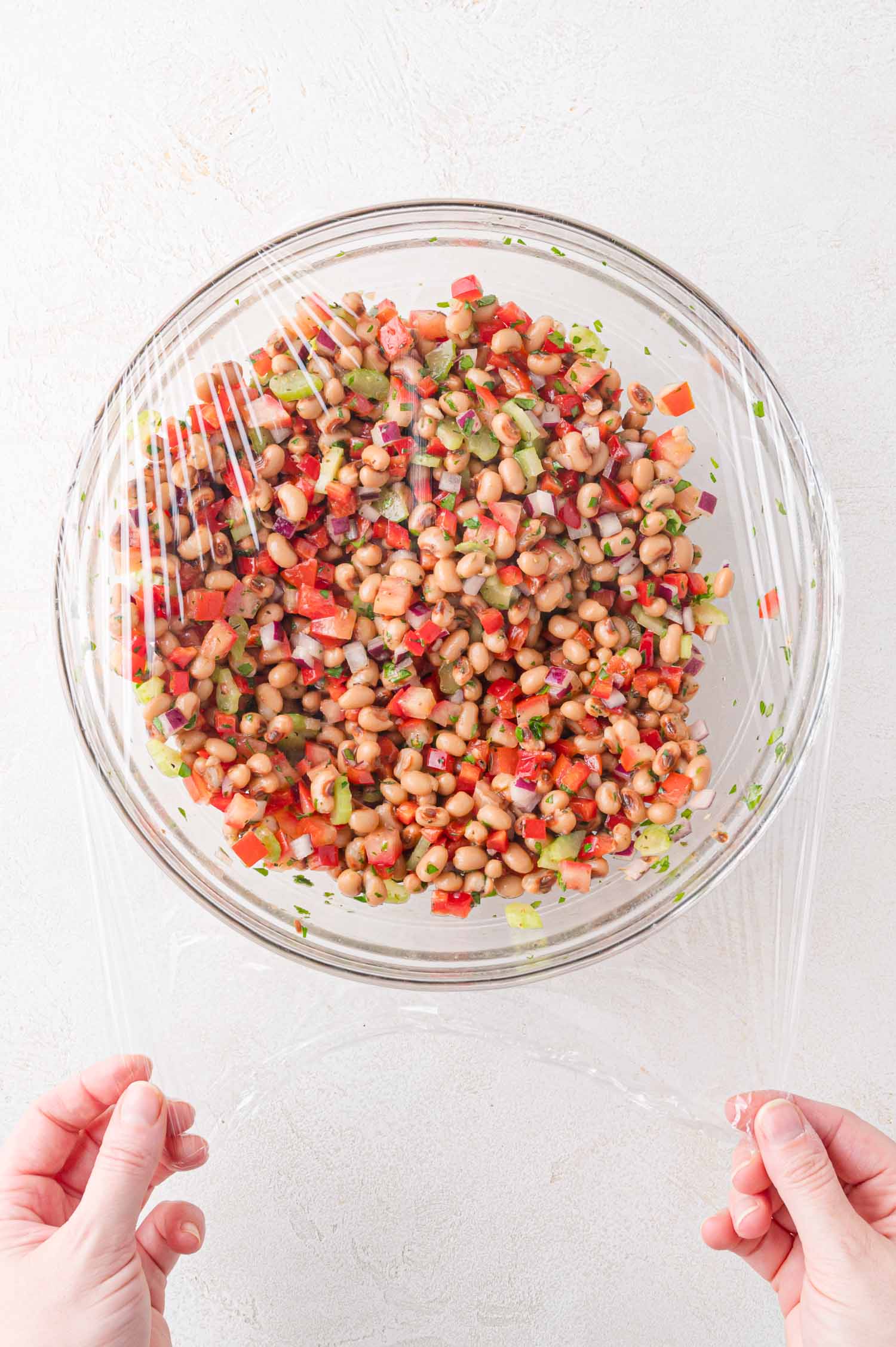 A person covers a glass bowl of black-eyed pea salad with plastic wrap on a white surface.