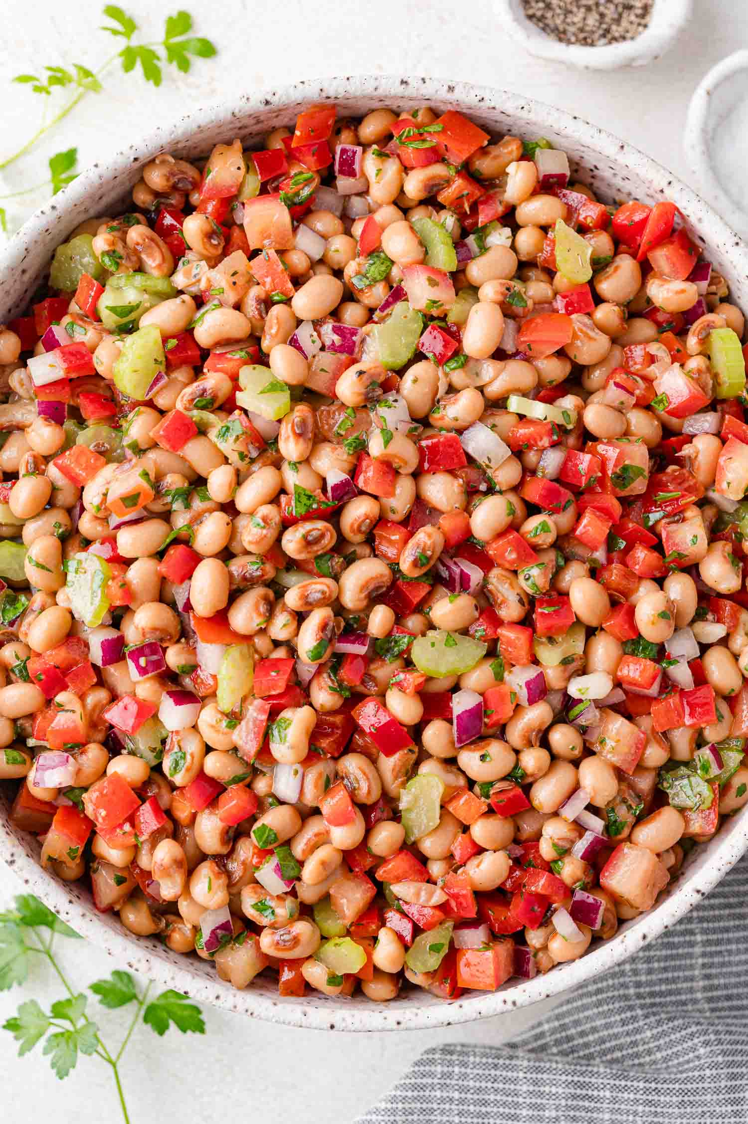 A bowl of black-eyed pea salad with diced red peppers, tomatoes, celery, onions, and herbs, served on a light surface with parsley garnish nearby.