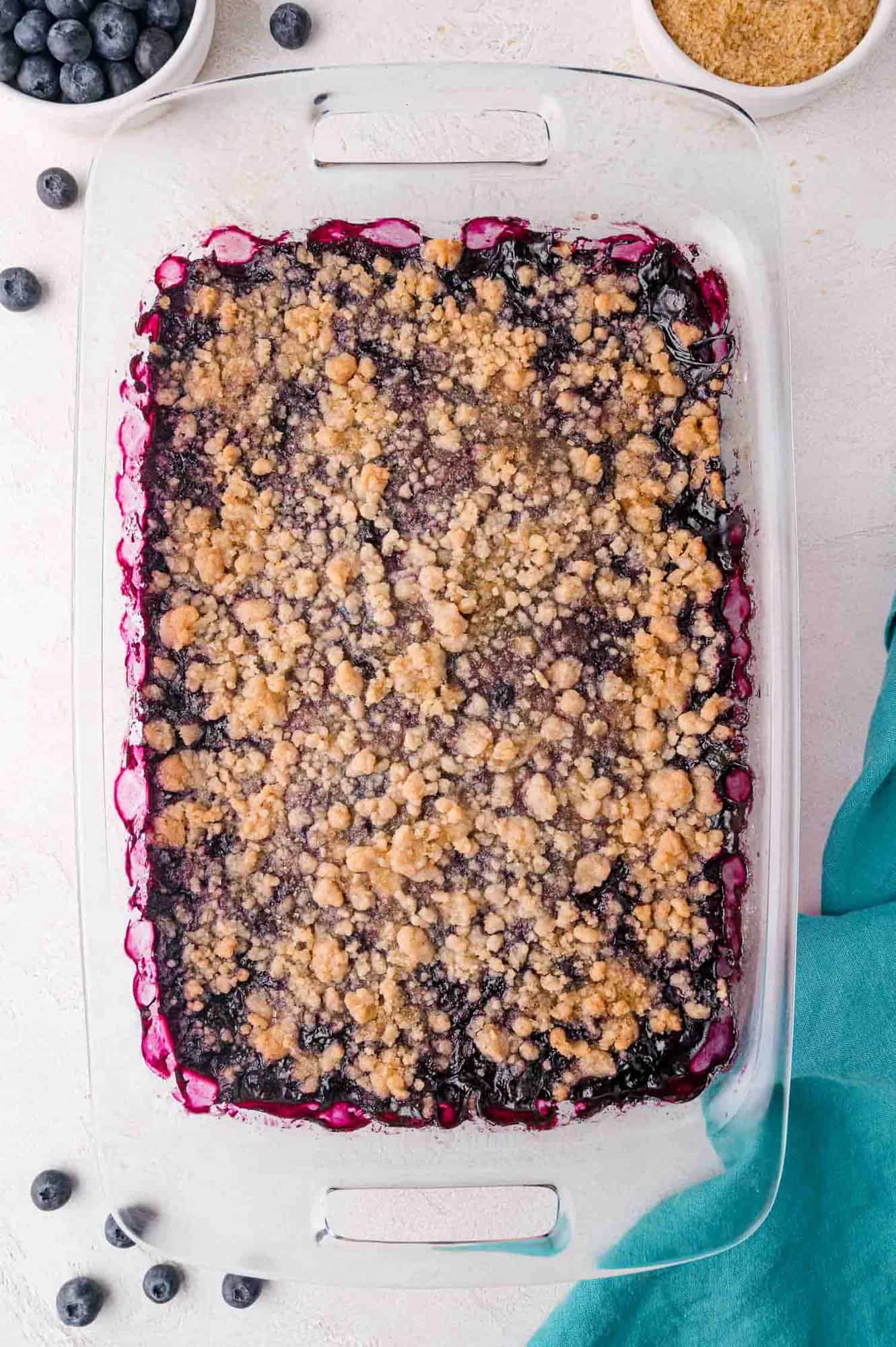 Blueberry Crumble 9 A glass baking dish filled with blueberry crumble, topped with a golden-brown streusel, sits on a light surface near a blue cloth and small bowls of blueberries and brown sugar.
