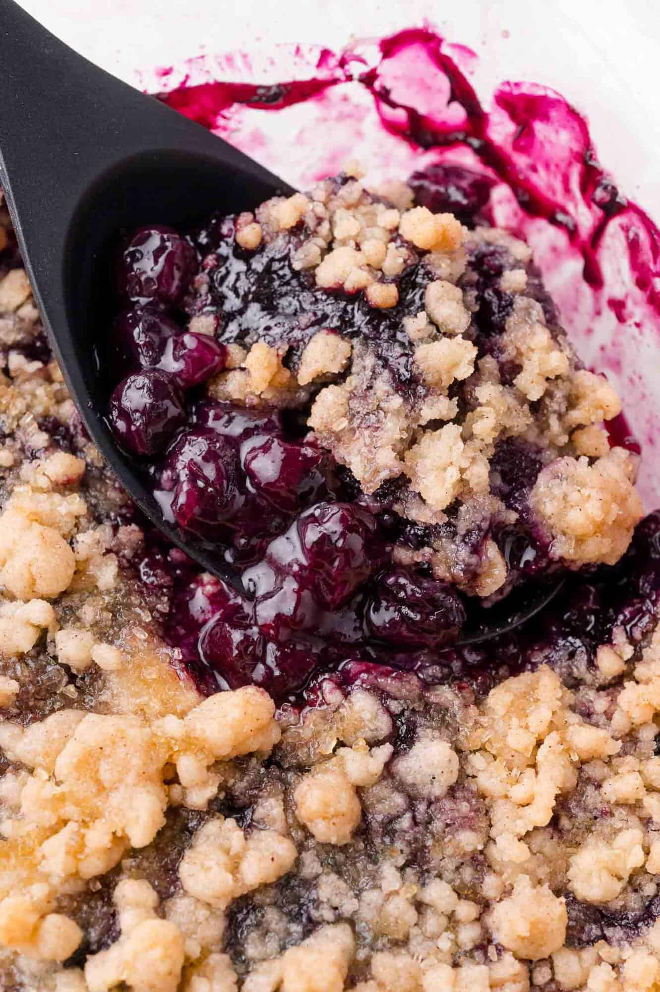 Blueberry Crumble 11 A close-up of a black spoon scooping blueberry crumble with a golden, crumbly topping and dark purple blueberry filling.