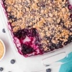 A glass baking dish filled with blueberry crumble, with one corner serving removed, showing the juicy blueberry filling and crumbly topping.