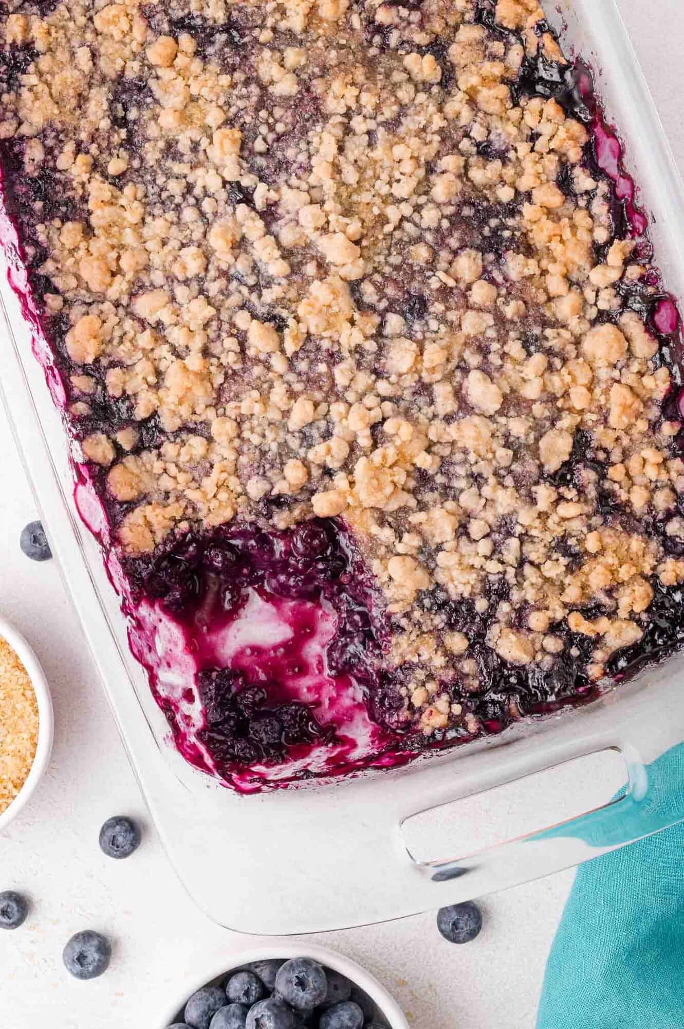 Blueberry Crumble 4 A baked blueberry crumble in a glass dish with a serving missing, surrounded by fresh blueberries and a bowl of brown sugar.