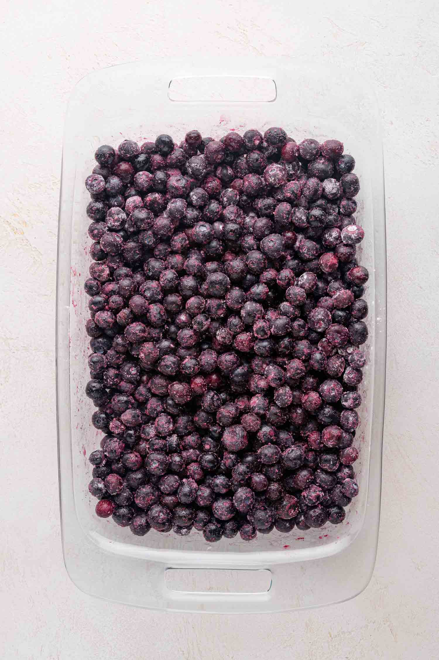 Blueberry Crumble 6 Glass baking dish filled with a layer of sugared blueberries on a light-colored surface.