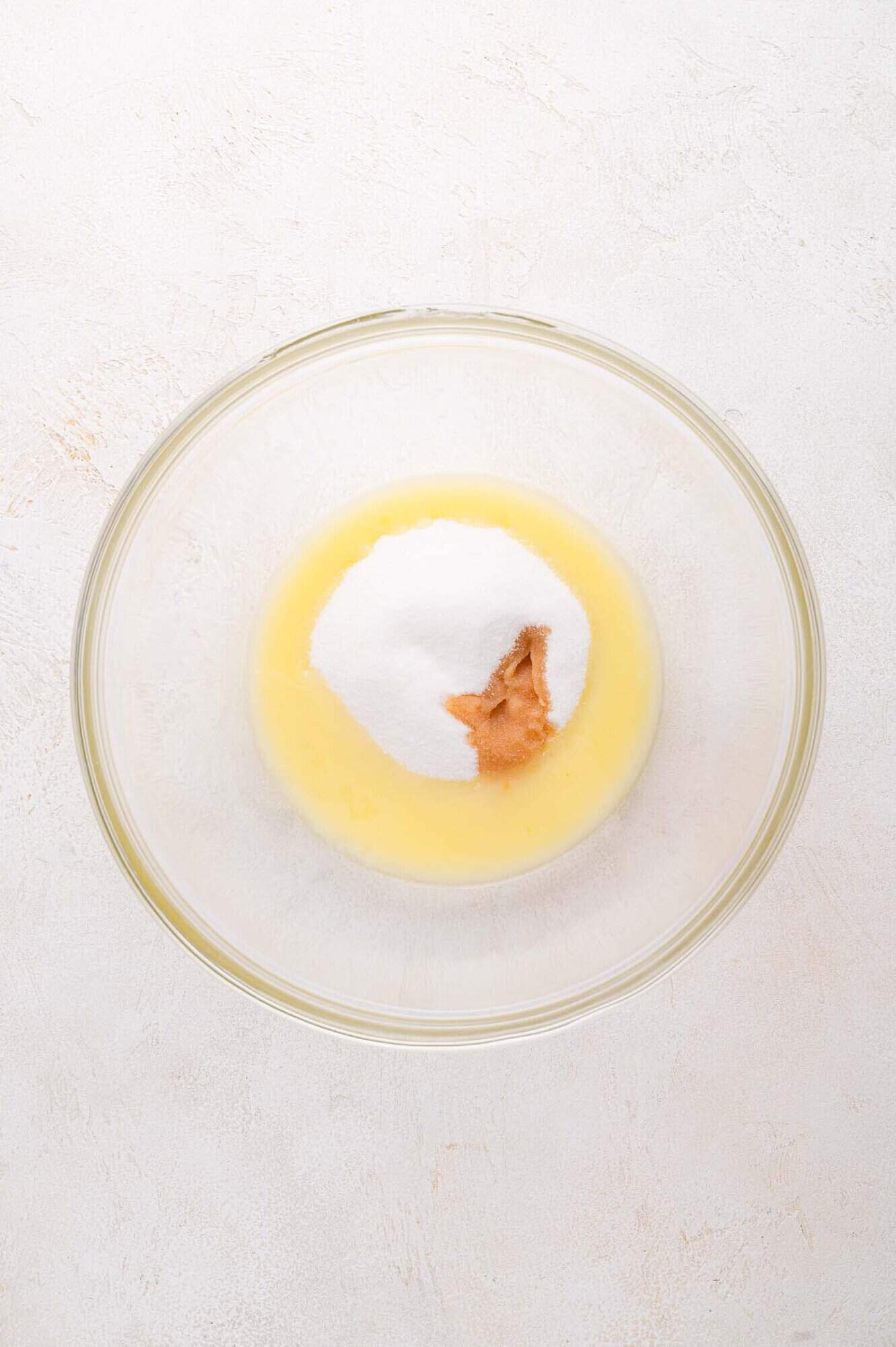 A glass mixing bowl containing melted butter, granulated sugar, and vanilla on a light-colored surface.