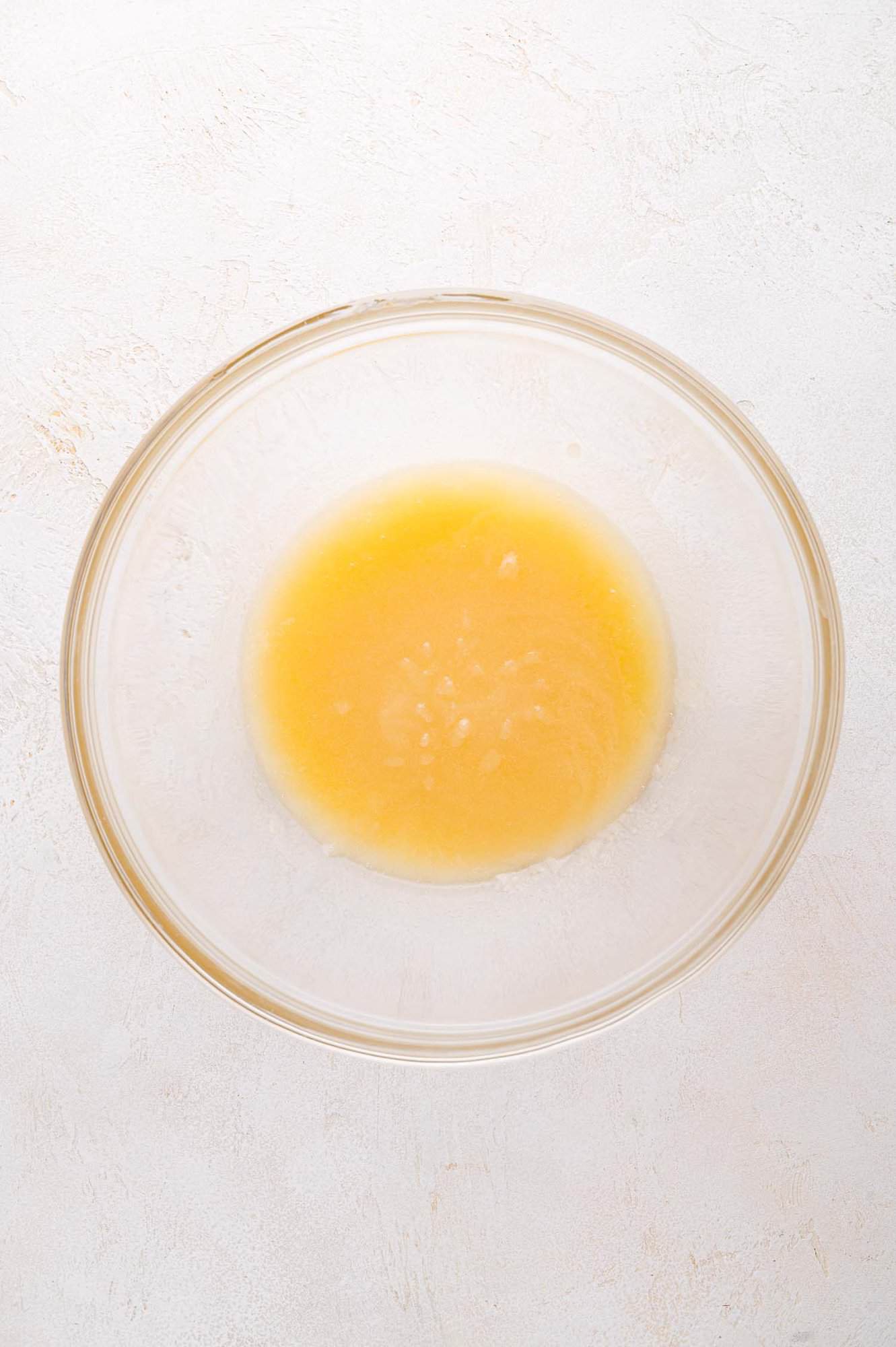 A glass bowl containing a small amount of yellow liquid mixture (butter, sugar, vanilla) sits on a light-colored surface.