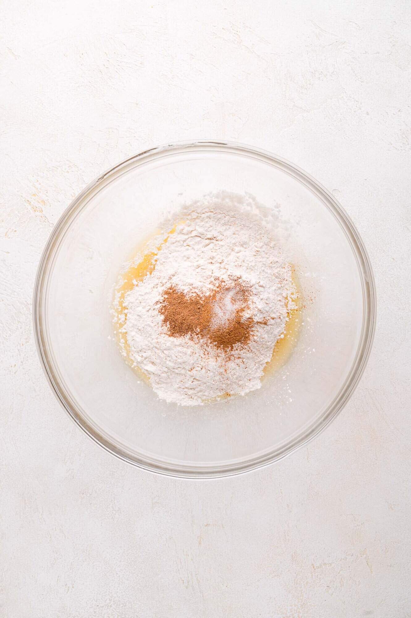 A glass bowl containing flour, cinnamon, and a wet ingredient mixture on a light-colored surface.