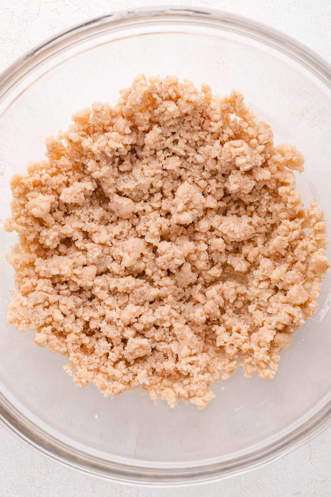 A clear glass bowl containing a crumbly, light brown mixture on a white surface.