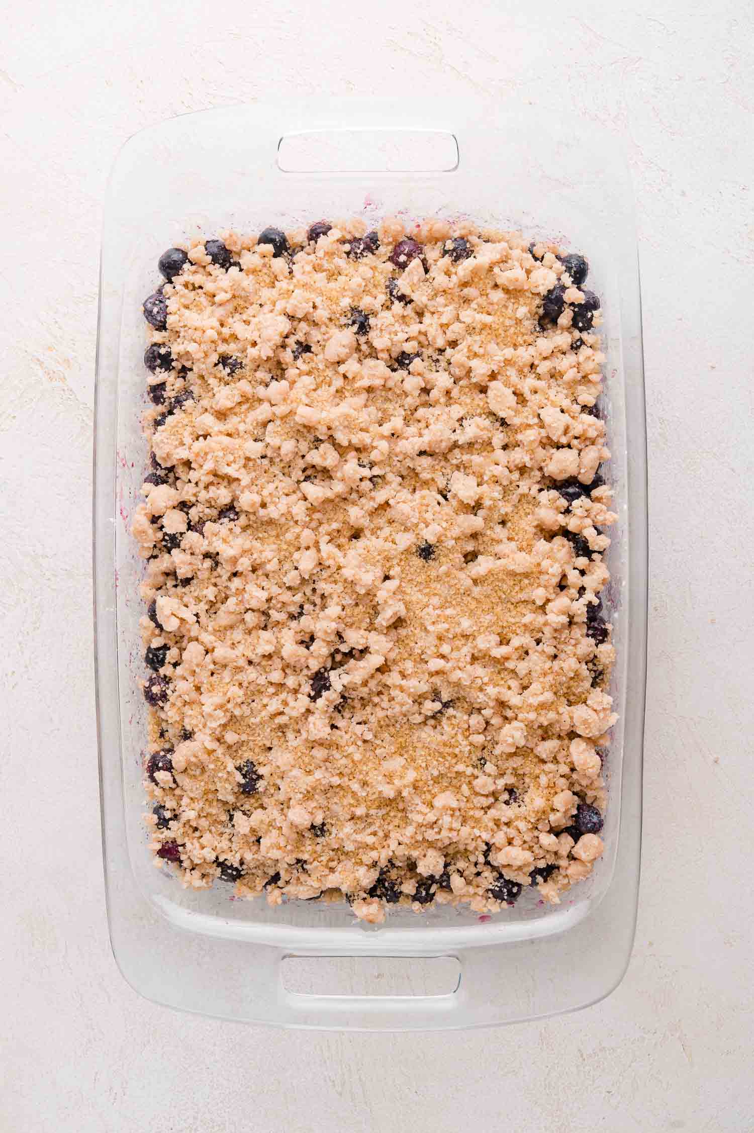 Blueberry Crumble 7 A rectangular glass baking dish filled with blueberry crumble topped with a layer of crumbly, golden-brown streusel, ready to be baked.