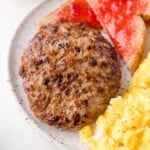 Two cooked sausage patties on a plate with scrambled eggs and toast topped with red jam.