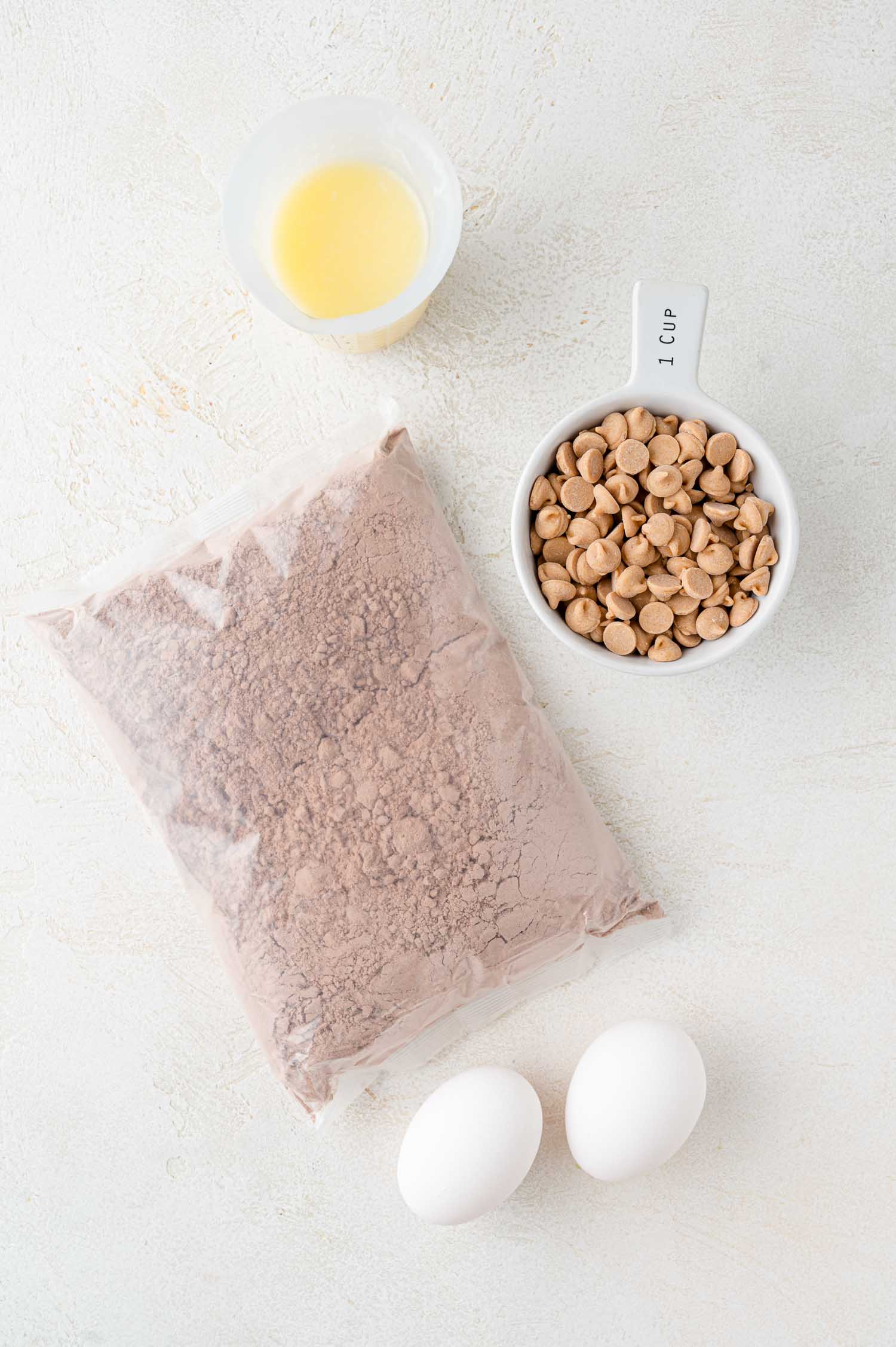 A bag of chocolate cake mix, two eggs, a cup of peanut butter chips, and a small cup of melted butter on a white surface.