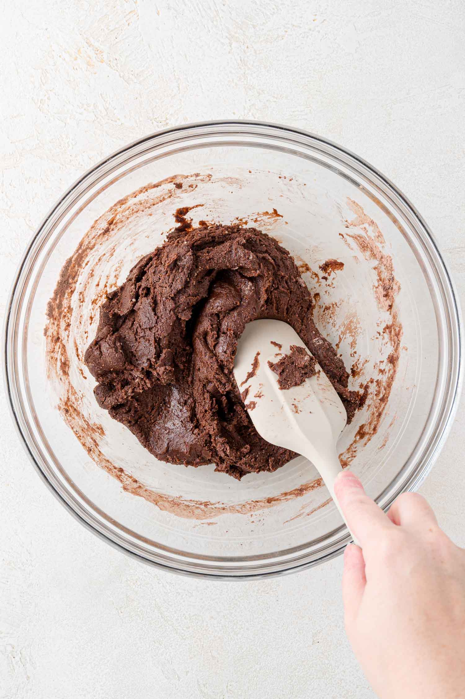 A hand holds a spatula mixing thick chocolate cookie dough in a clear glass bowl on a light-colored surface.