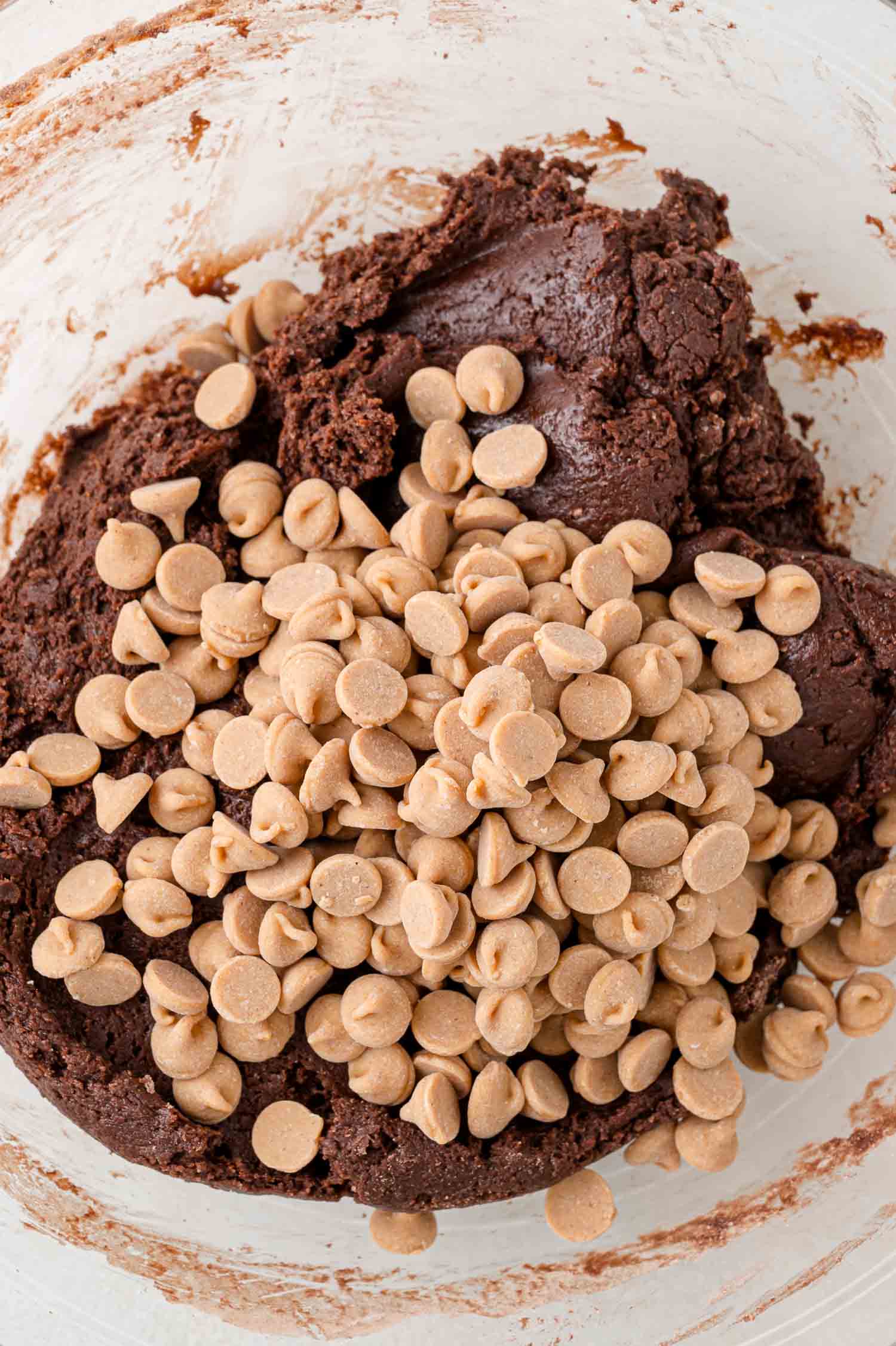 A mixing bowl with chocolate cookie dough topped with a pile of peanut butter chips, ready to be combined.