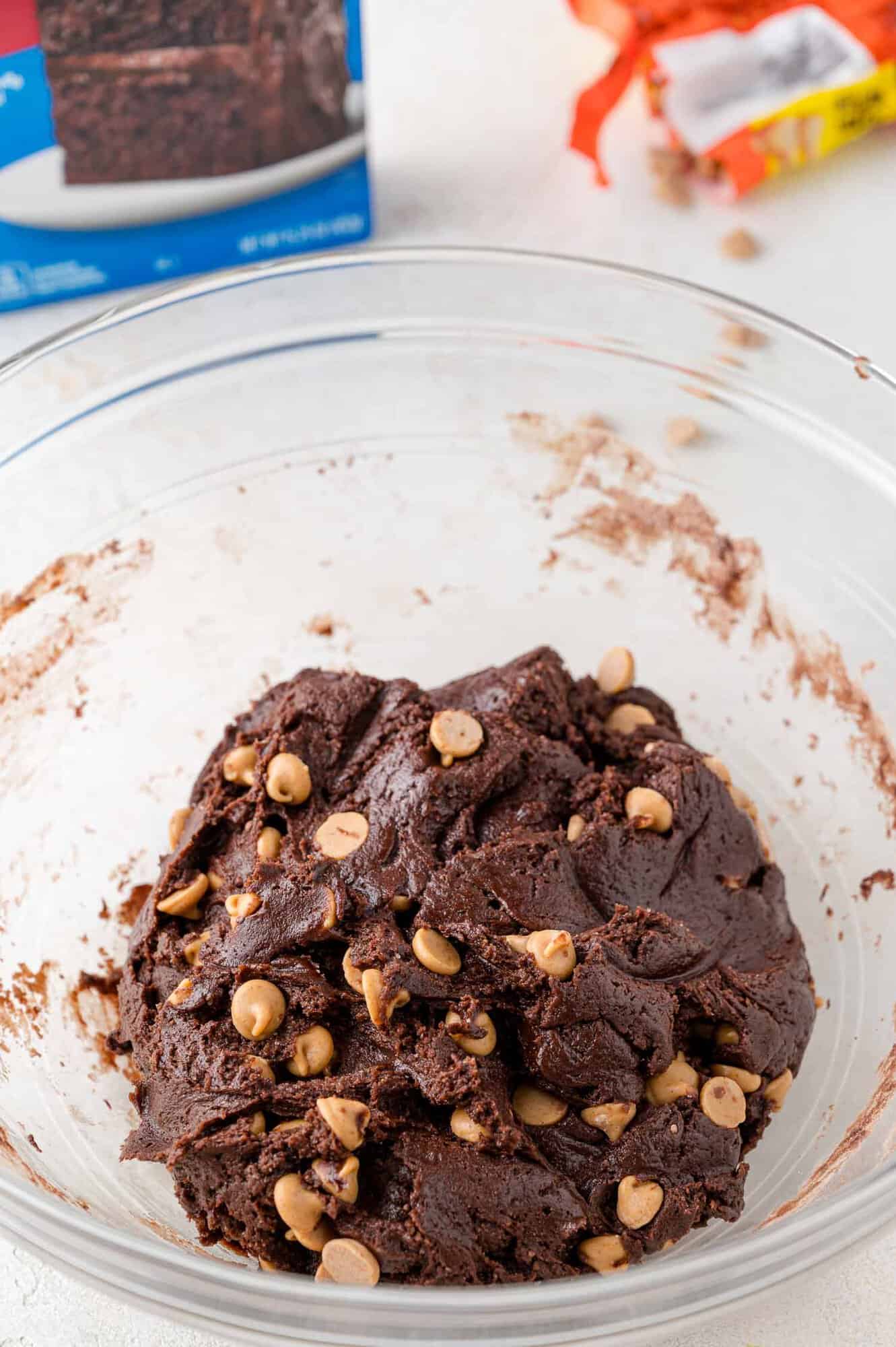 A glass bowl filled with chocolate cookie dough mixed with peanut butter chips sits on a light surface; a box and candy wrapper are in the background.