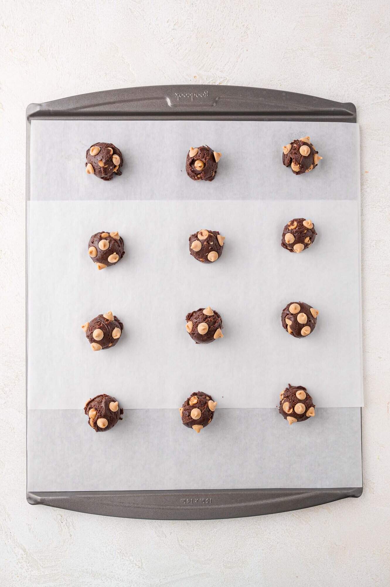A baking sheet lined with parchment paper holds twelve evenly spaced scoops of chocolate cookie dough with white chocolate chips.