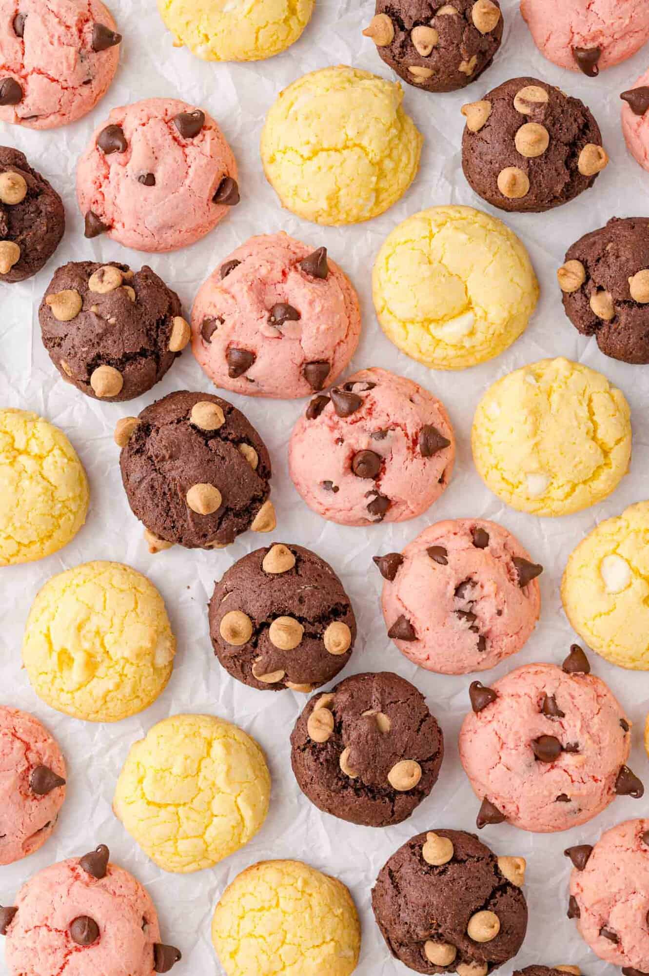 Rows of pink, yellow, and brown cookies, some topped with chocolate chips, arranged on a white parchment paper background.