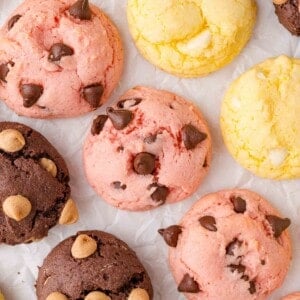 Assorted cake mix cookies with chocolate chips and white chocolate chips in pink, yellow, and brown colors are arranged on crumpled white parchment paper.