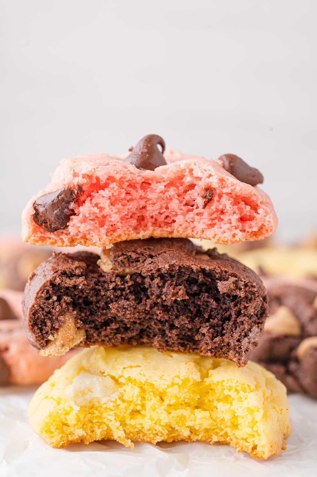 Three thick cookies—pink with chocolate chips, chocolate, and yellow—are stacked, each with a bite taken out of them.
