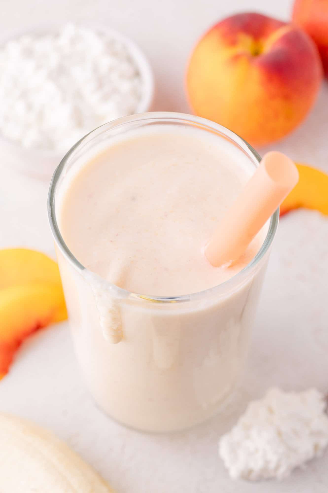 Cottage Cheese Smoothie 4 A glass of creamy smoothie with a straw, surrounded by peach slices, a whole peach, cottage cheese, and a banana on a white surface.
