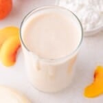 A glass of creamy peach cottage cheese smoothie sits on a white surface, with peach slices and a bowl of cottage cheese beside it.