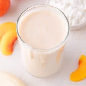 A glass of creamy peach cottage cheese smoothie sits on a white surface, with peach slices and a bowl of cottage cheese beside it.
