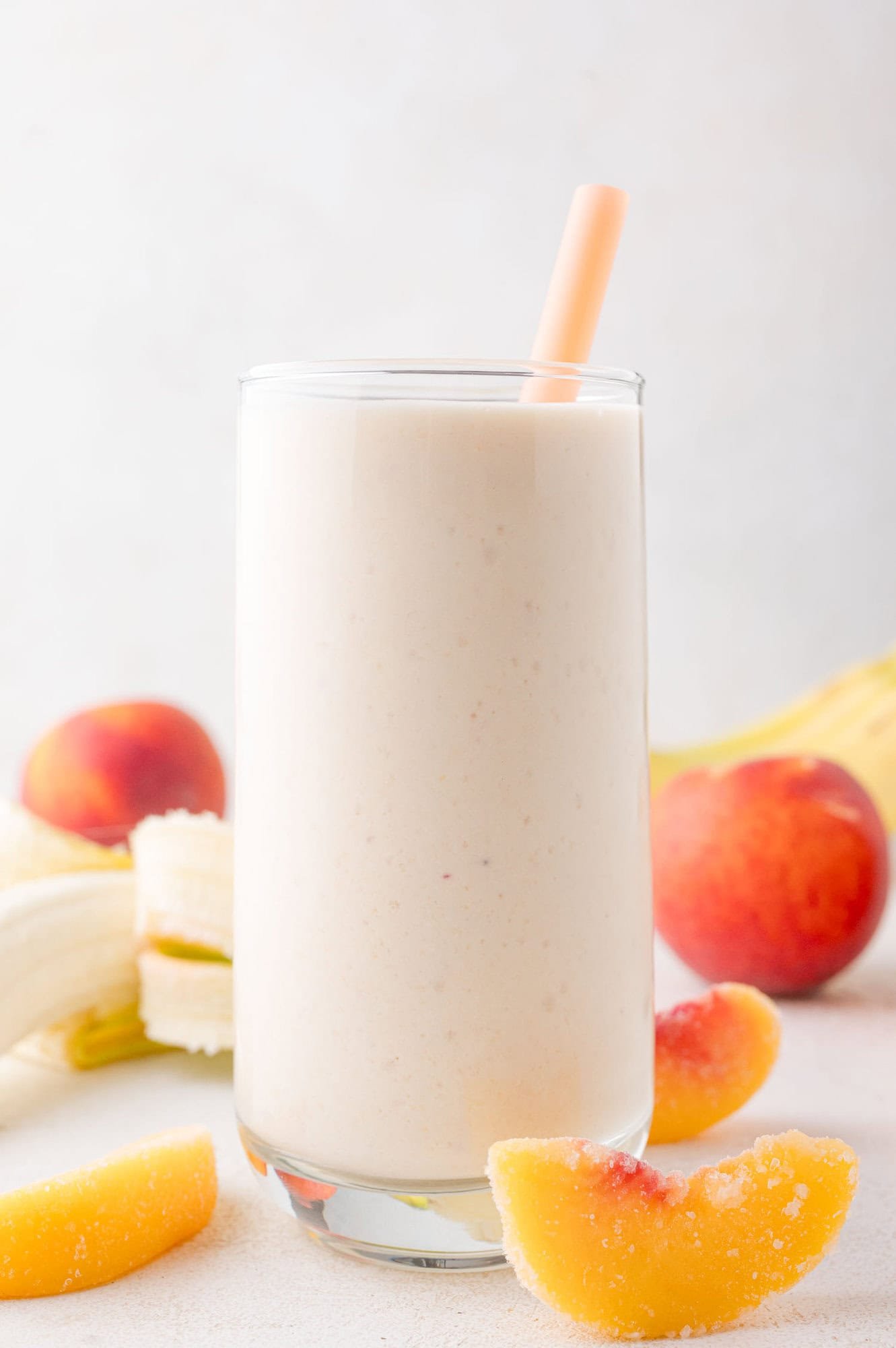 Cottage Cheese Smoothie 2 A glass of creamy smoothie with a straw, surrounded by peach slices, bananas, and whole peaches on a light background.