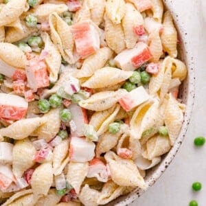 A close-up of a pasta salad with shell pasta, imitation crab meat, green peas, and diced red onion in a creamy dressing.